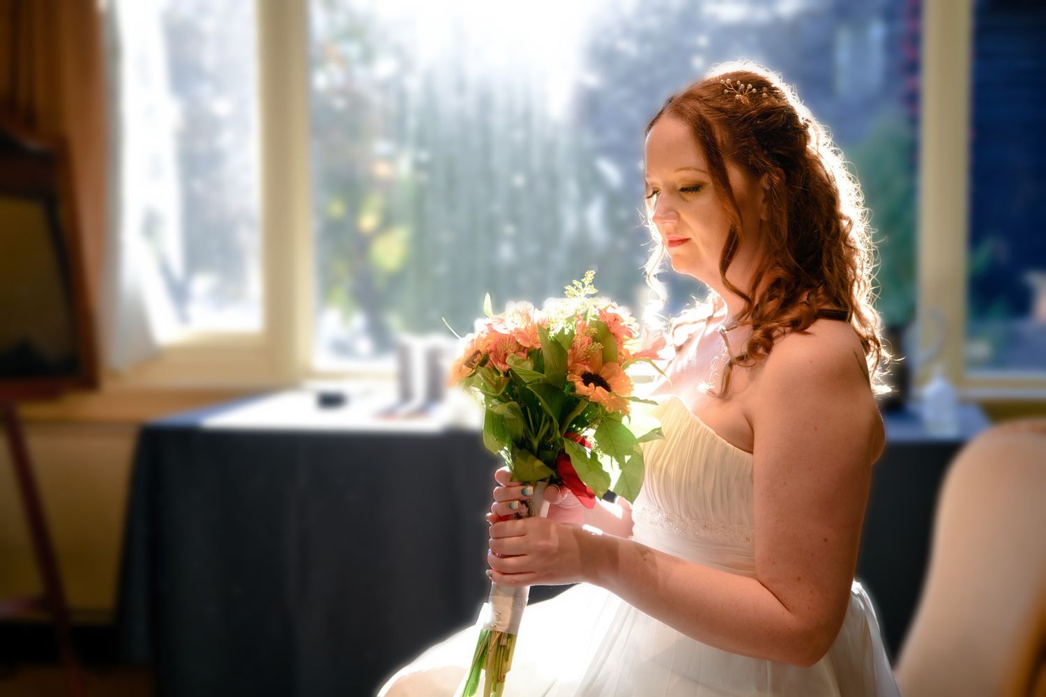 A bride in an elegant strapless gown holds a delicate bouquet while standing in a bright sunlit room.