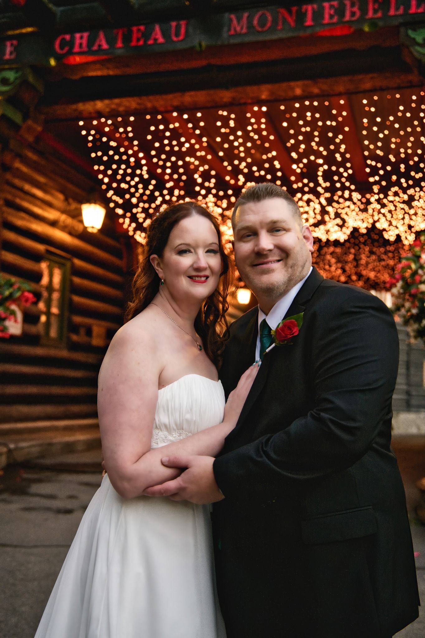 A romantic wedding couple poses under twinkling lights at a rustic venue in their formal attire.