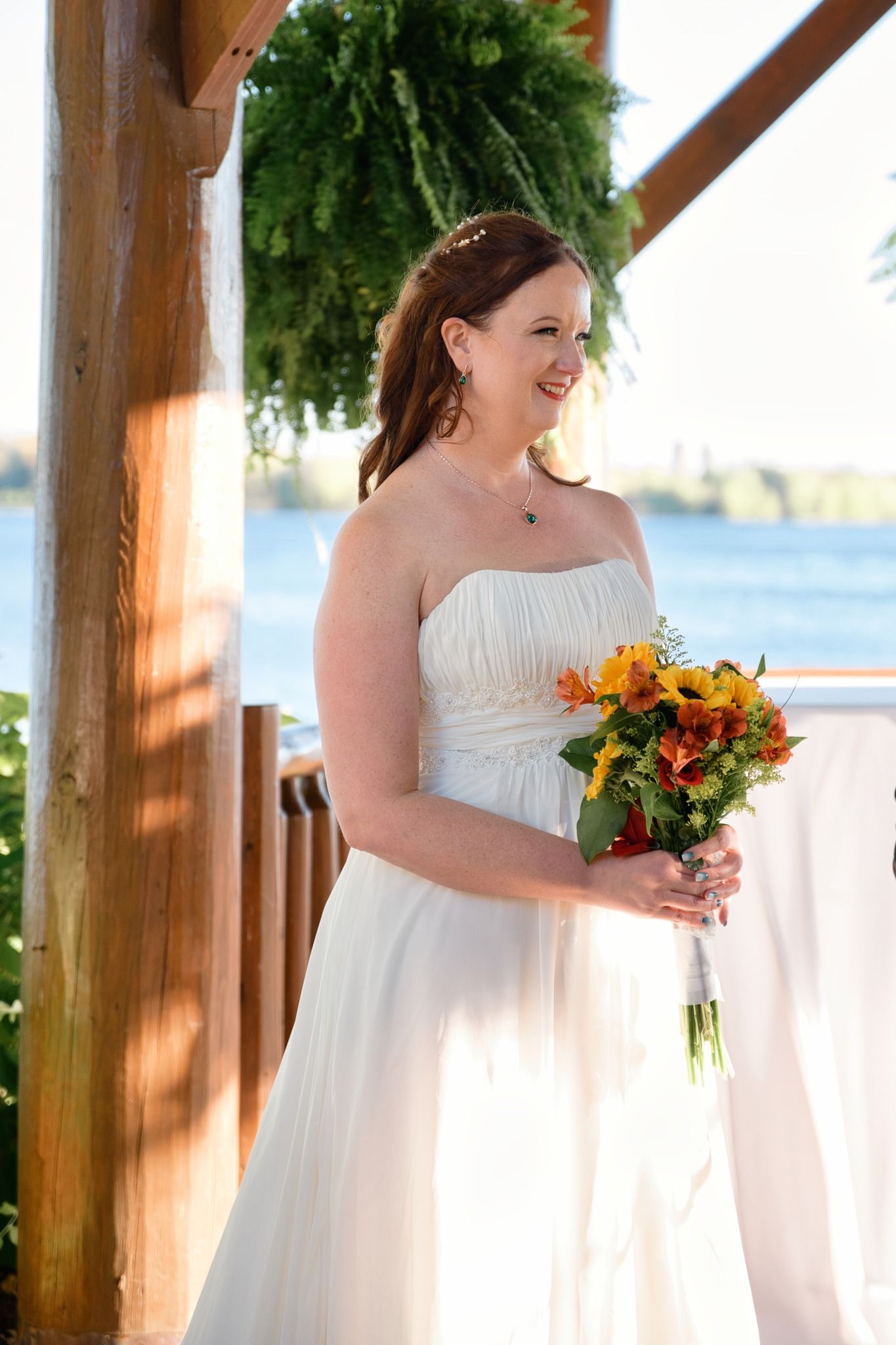 A bride in a white dress stands among hanging ferns holding an orange and yellow bouquet.