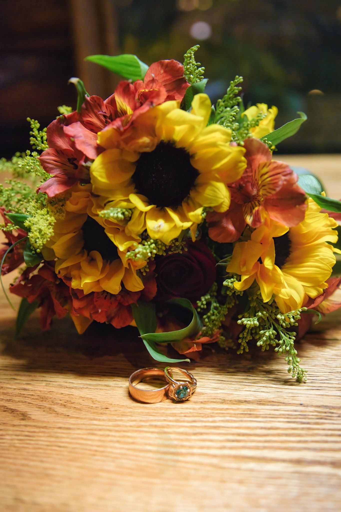 Vibrant autumn wedding bouquet with sunflowers and fall colors sits next to wedding rings on wooden surface.