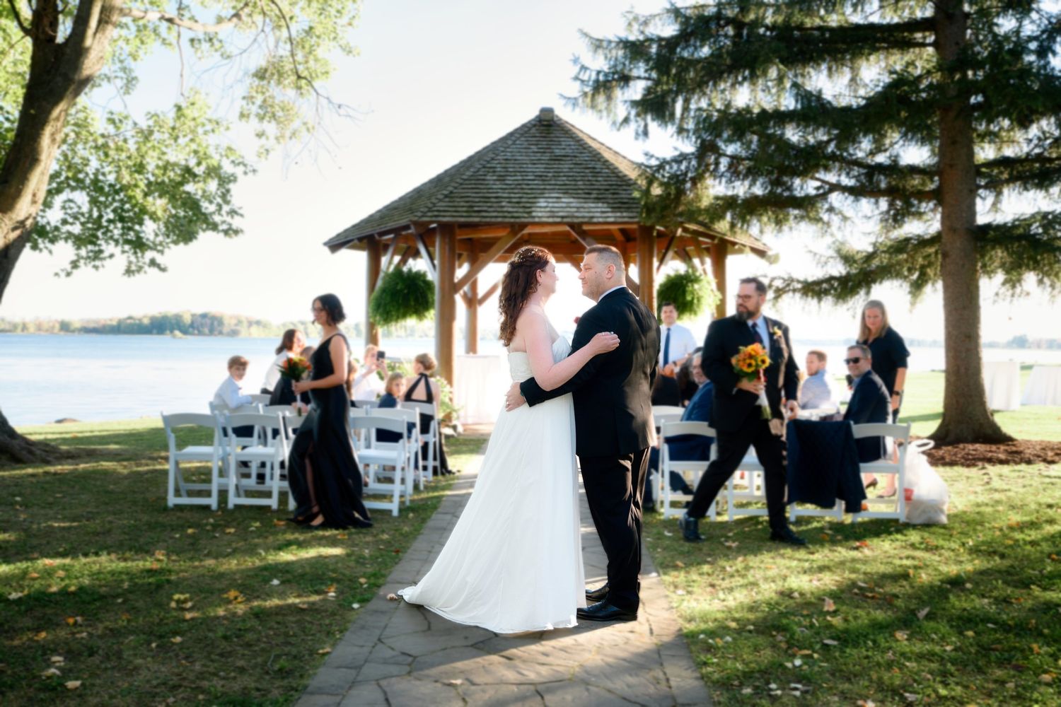 Wedding ceremony setup along a serene lakefront with white chairs and gazebo under mature trees in golden afternoon light.