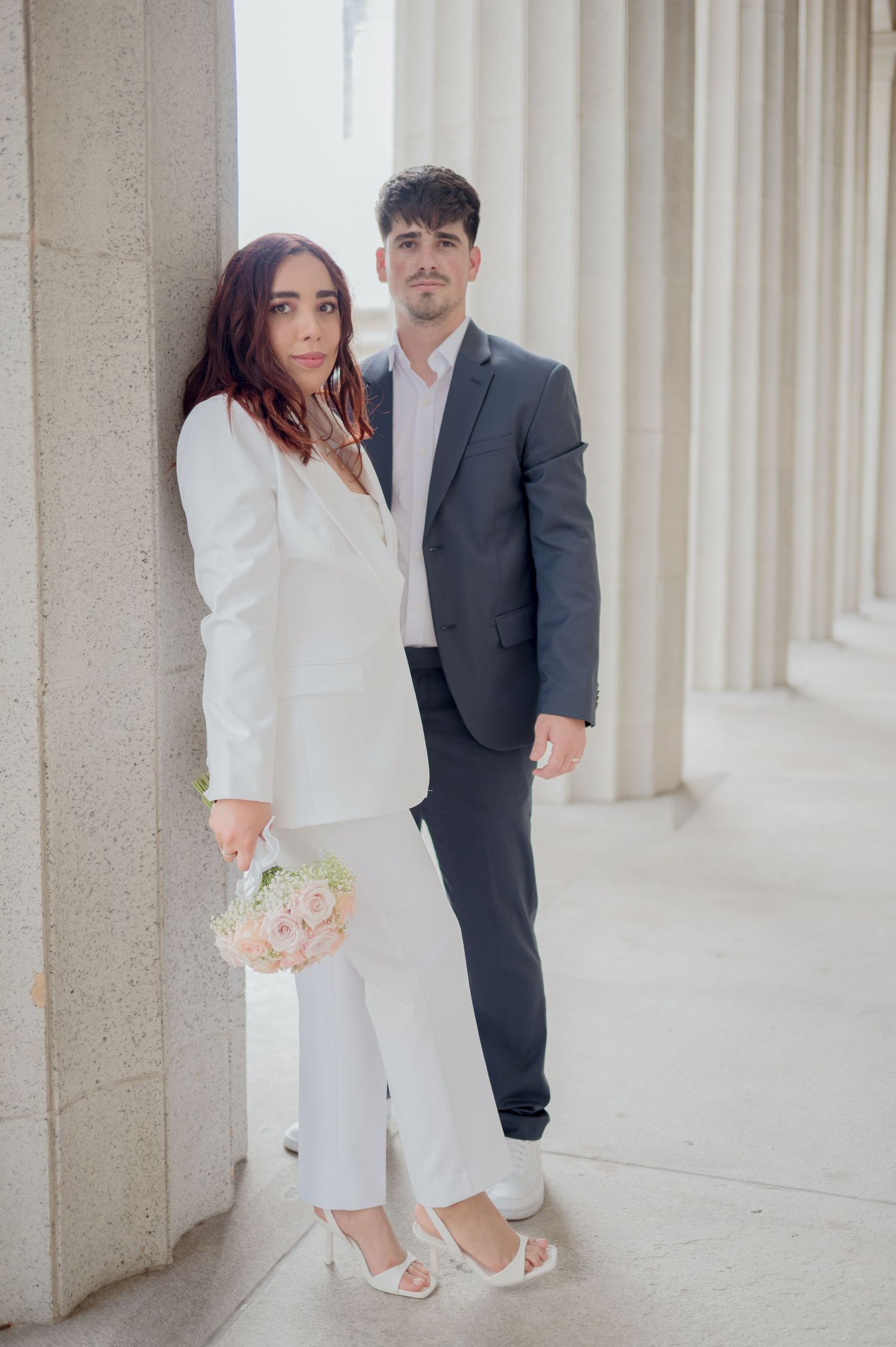 Elegant couple in white and navy attire poses against stone columns in romantic wedding photography series.