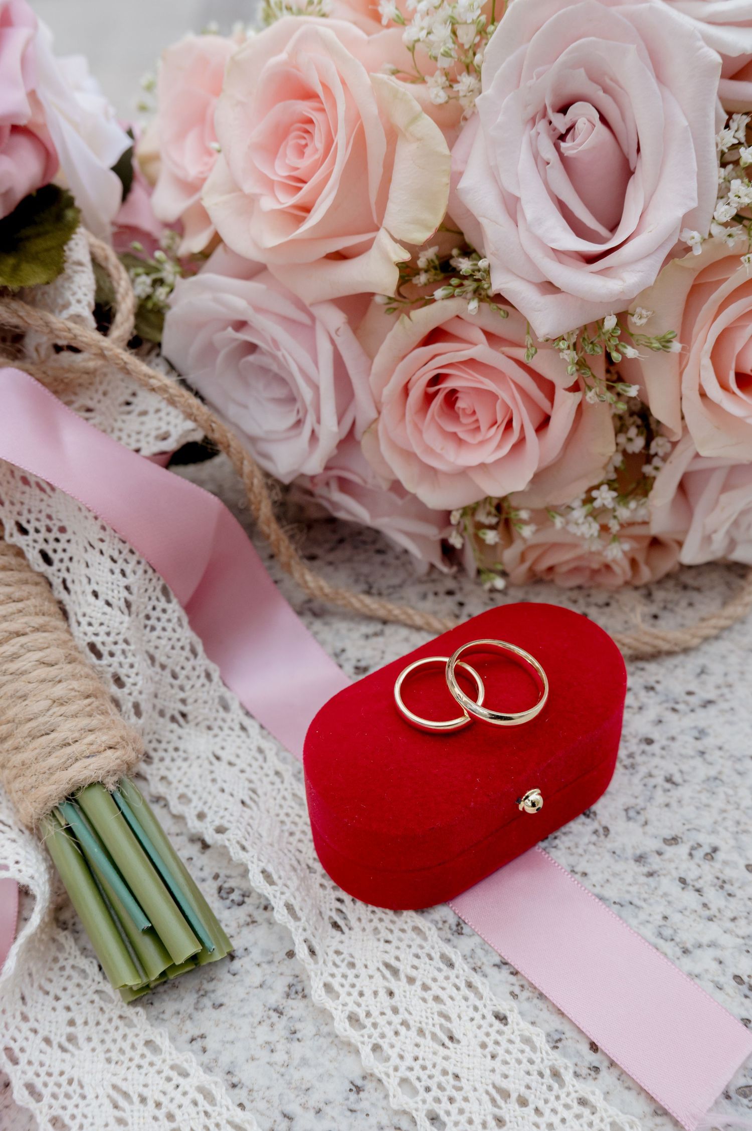Pink roses and baby's breath wedding bouquet with gold wedding bands resting on red velvet heart cushion and lace backdrop.