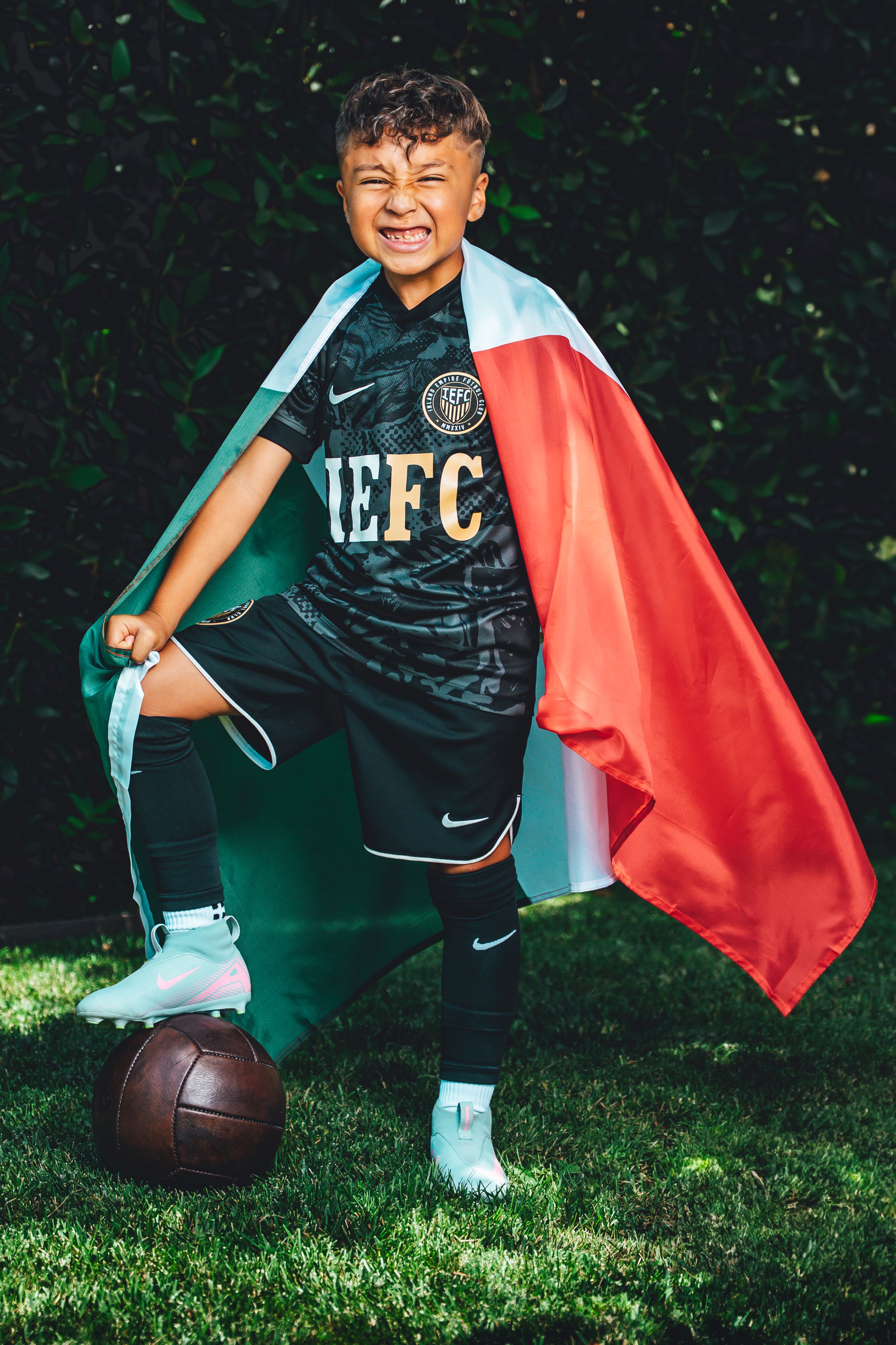 Young athlete in black jersey poses with football and red cape against green backdrop.