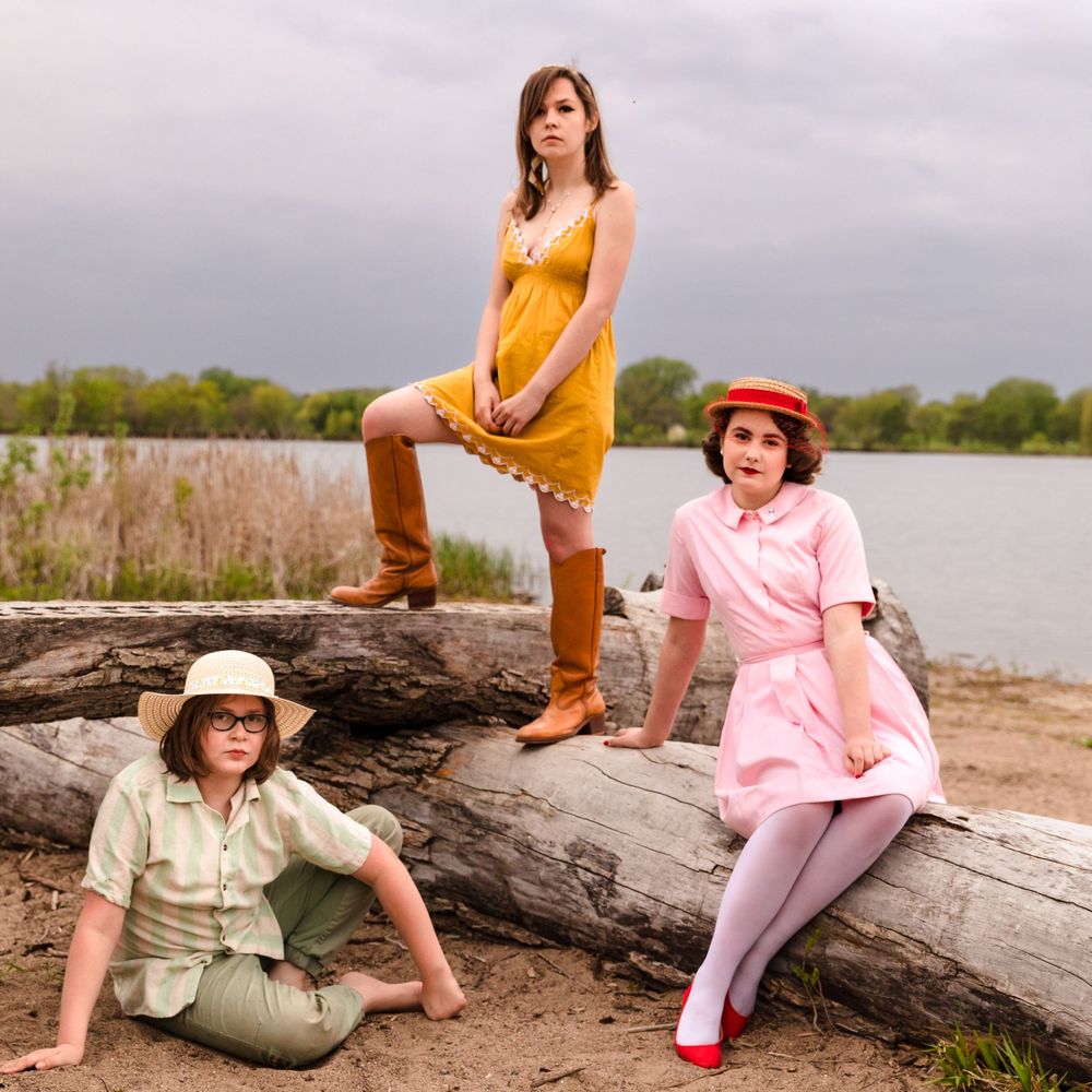 Three people in vintage-style outfits pose on a fallen log by a lake shore during an overcast day.