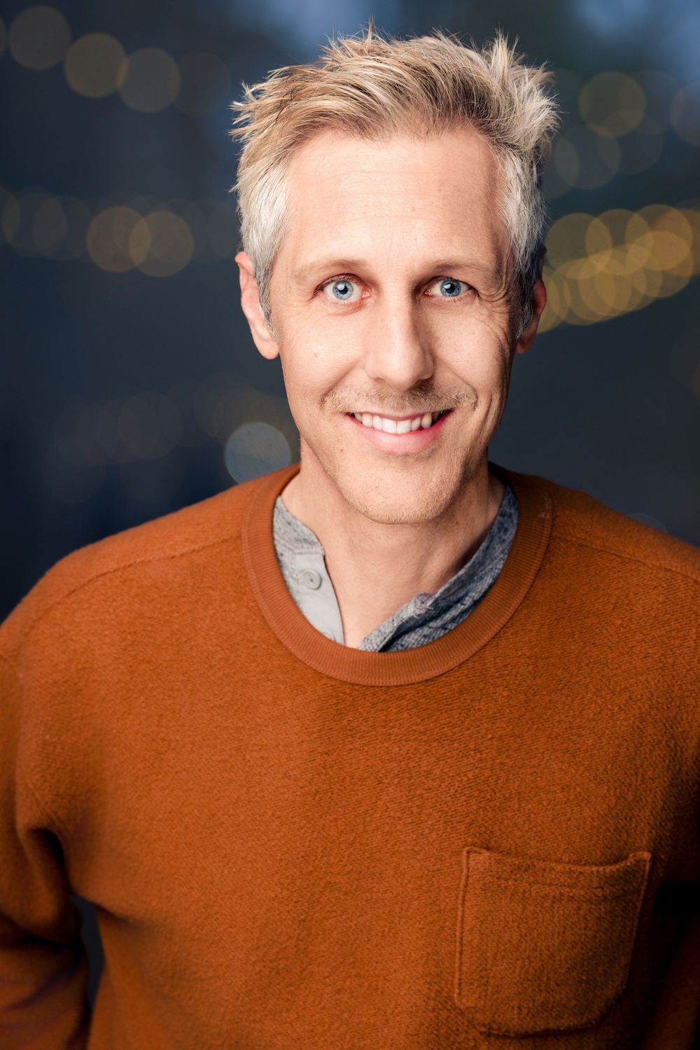 Professional portrait of someone with silver hair wearing a rust-colored sweater with a grey collar against a blurred background.