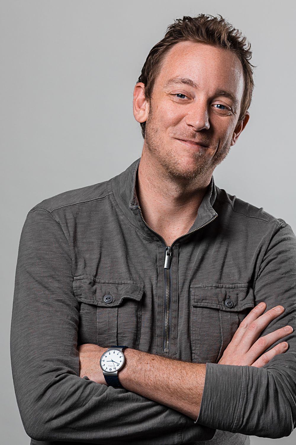 Actor headshot of person in gray zip-up jacket with rolled sleeves wearing watch against light background. Mika Larson Photography.