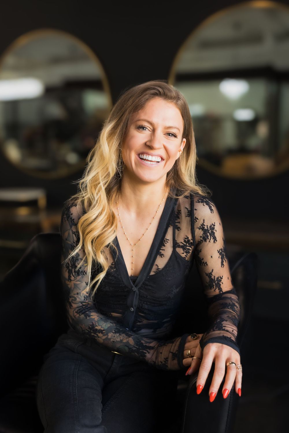 Woman in black lace top and blonde hair smiles while sitting in dimly lit restaurant setting.
