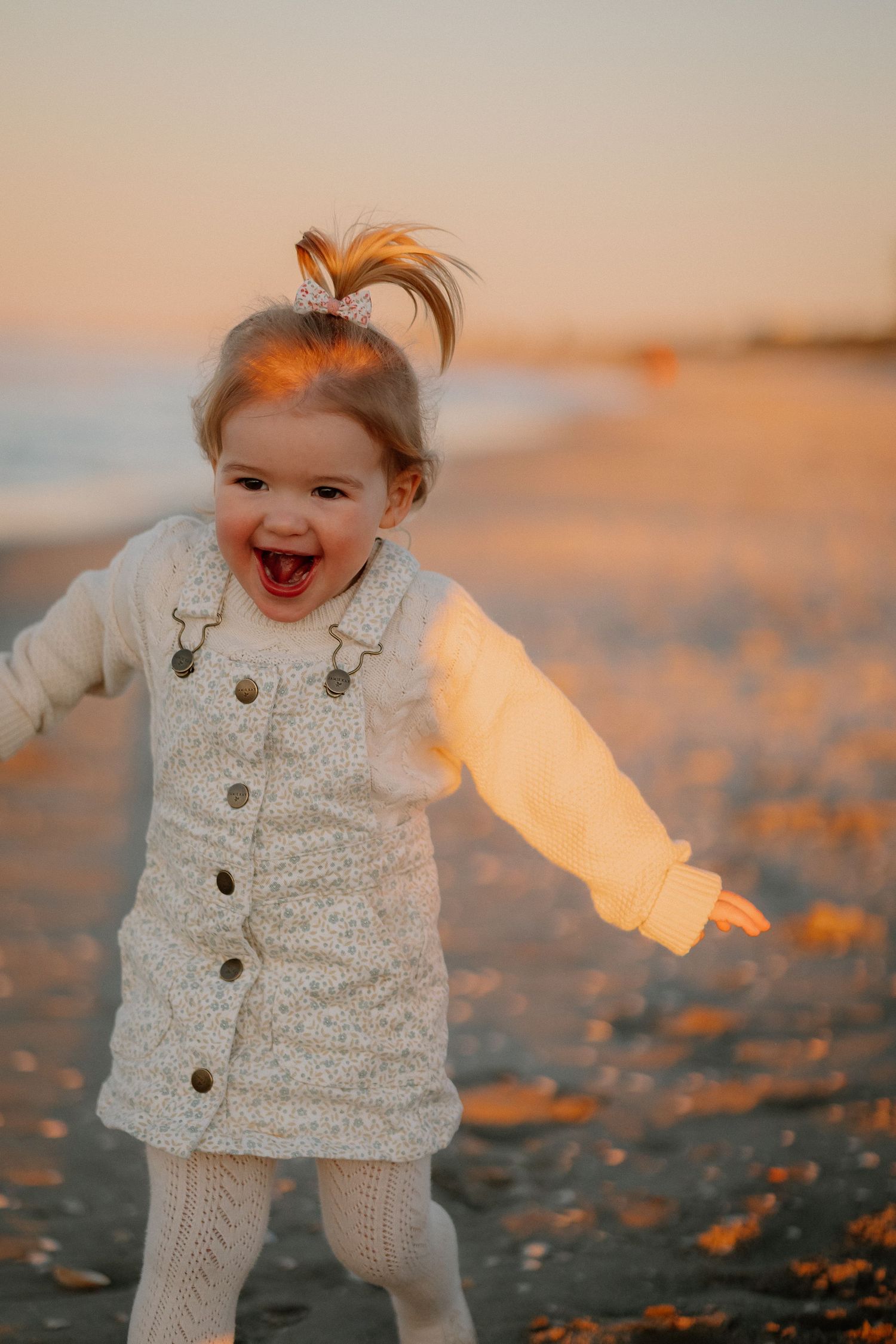 A child in a cozy white knit sweater and tights plays joyfully among autumn leaves at sunset.