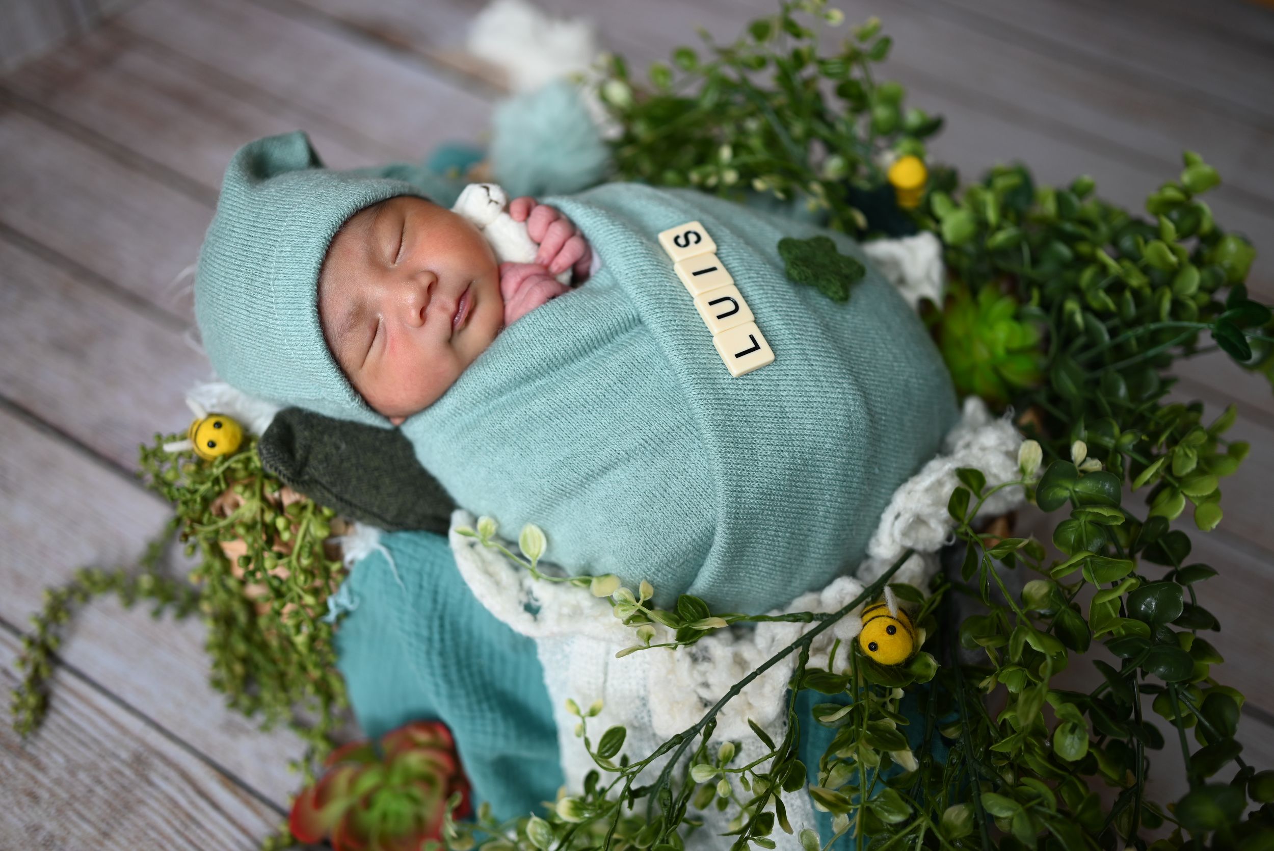 A sleeping baby swaddled in teal fabric lies on a rustic wooden surface surrounded by fresh greenery and white flowers.