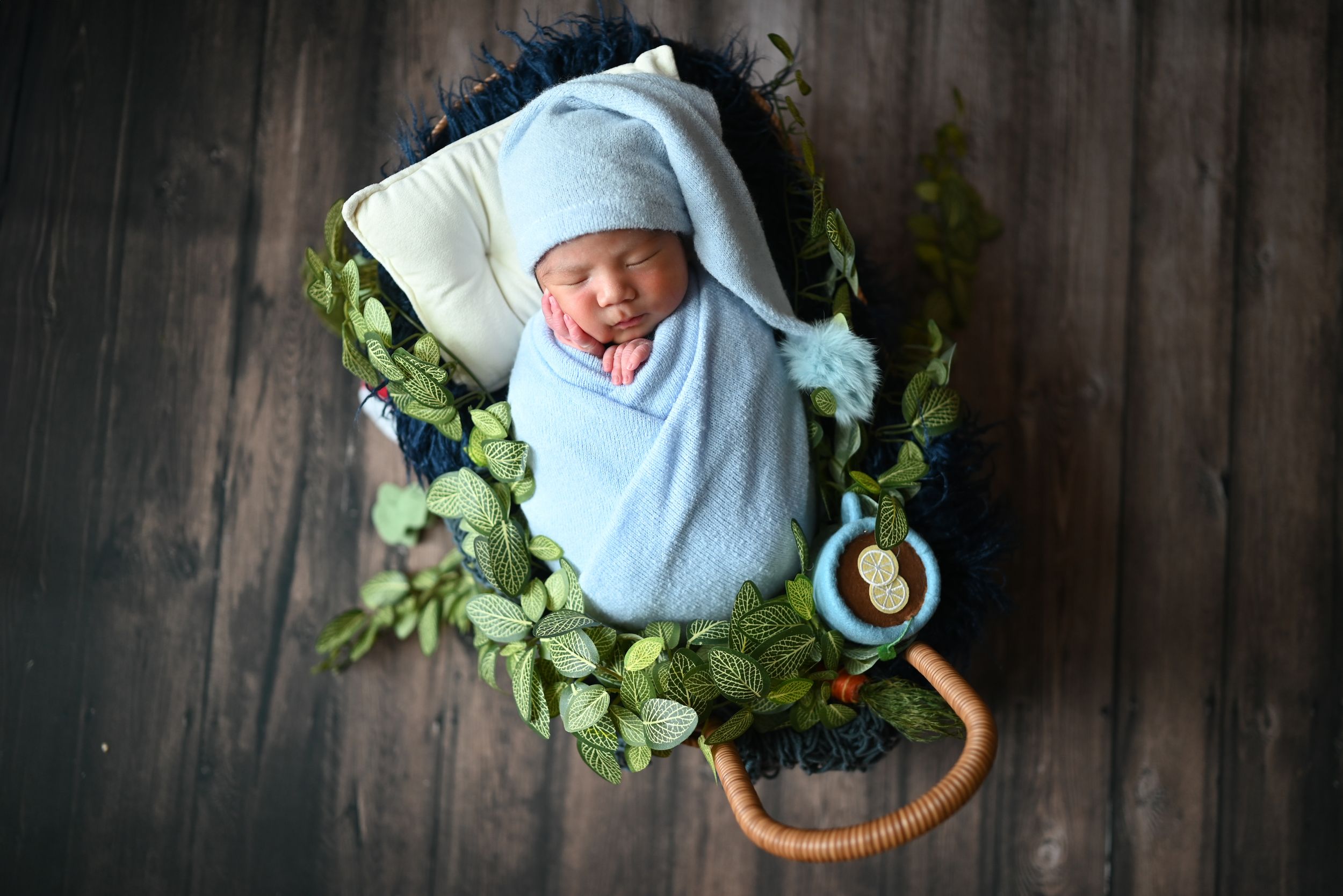Sleeping newborn wrapped in light blue blanket nestled in basket with greenery on dark wooden floor.