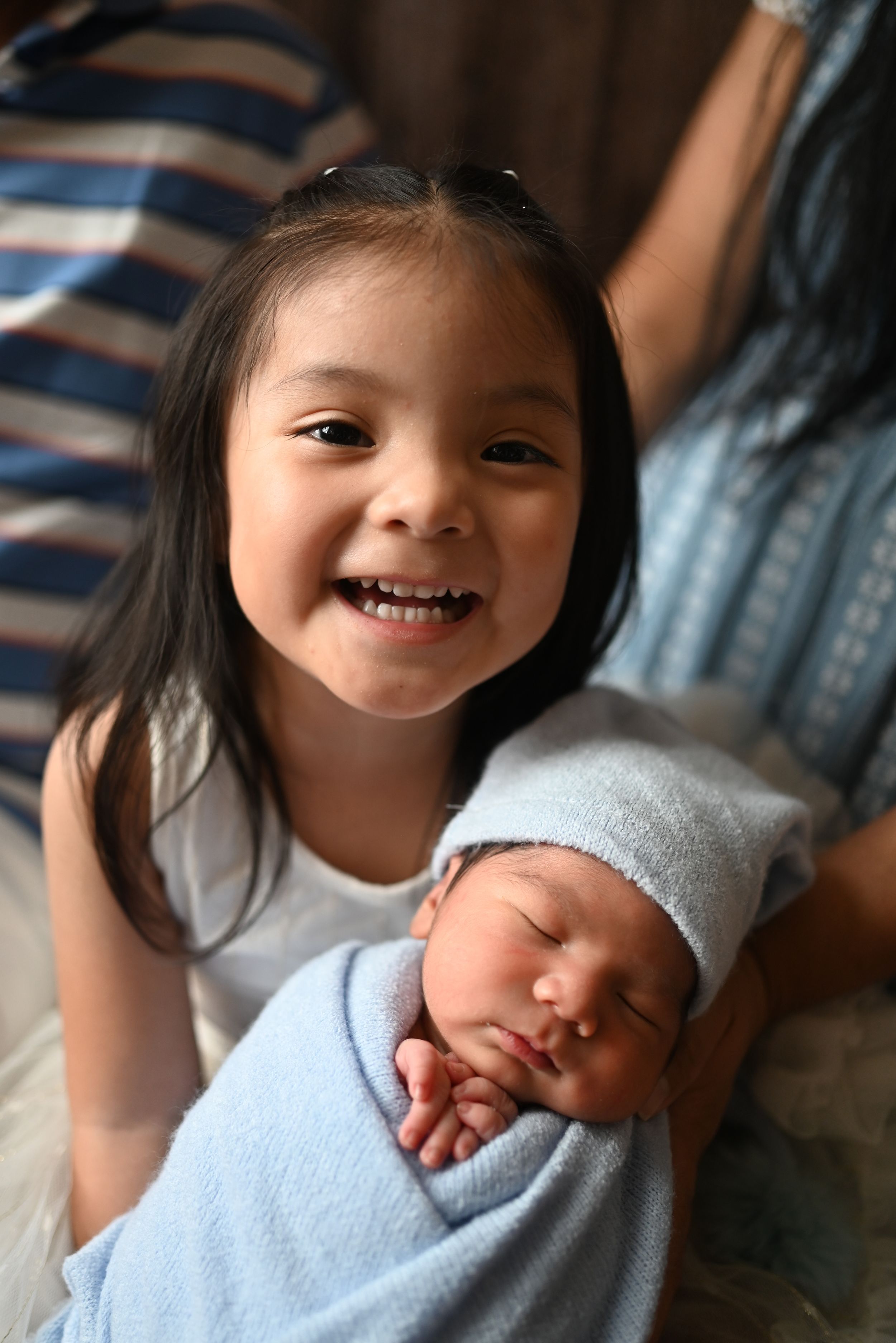 A joyful sibling moment as a young sister poses with a sleeping newborn baby in blue.
