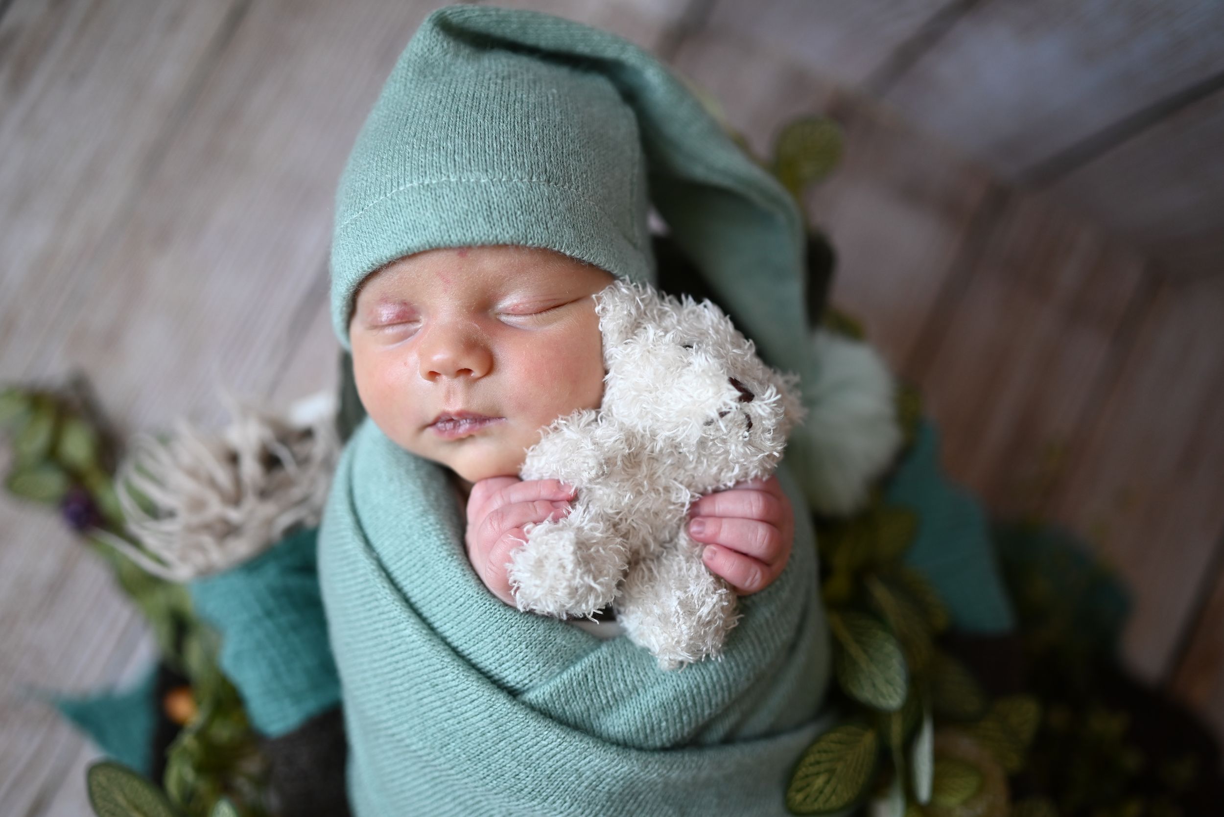 A sleeping baby swaddled in a mint green wrap with a small stuffed lamb toy.