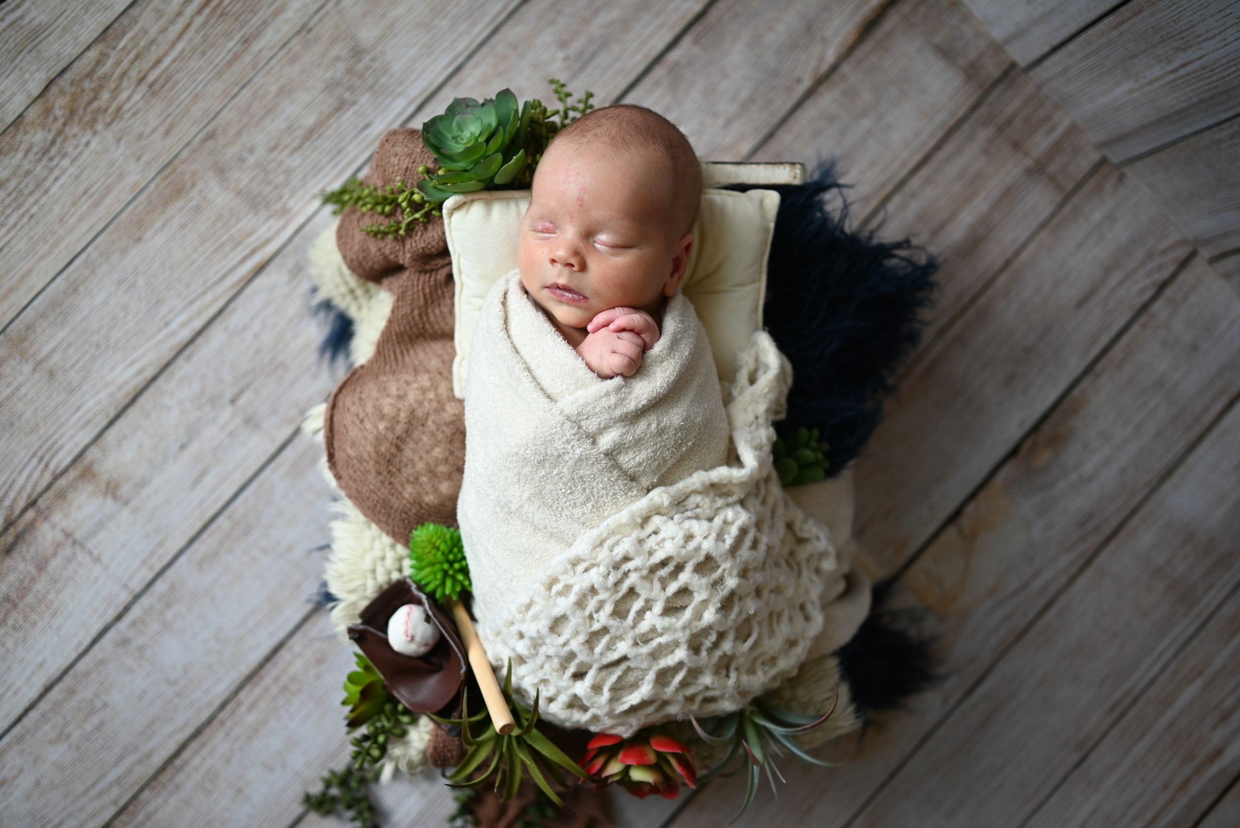 Baby in white knit outfit lies on rustic wooden floor with greenery and floral decorative accents.