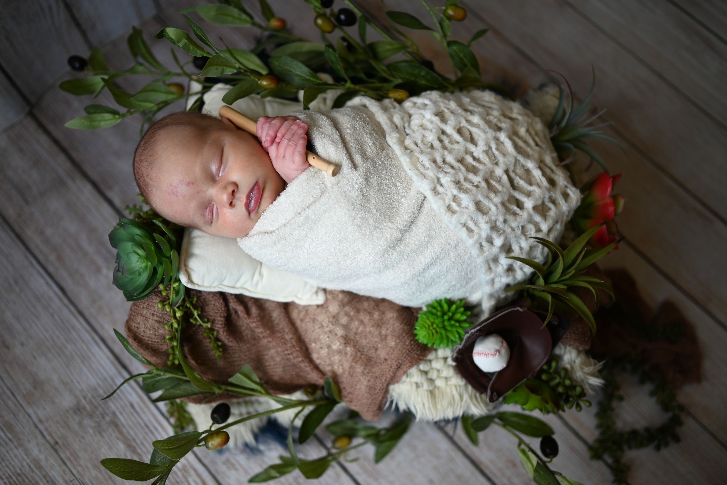 Baby wearing white dress lying on rustic wooden floor with scattered greenery and red floral decorations.