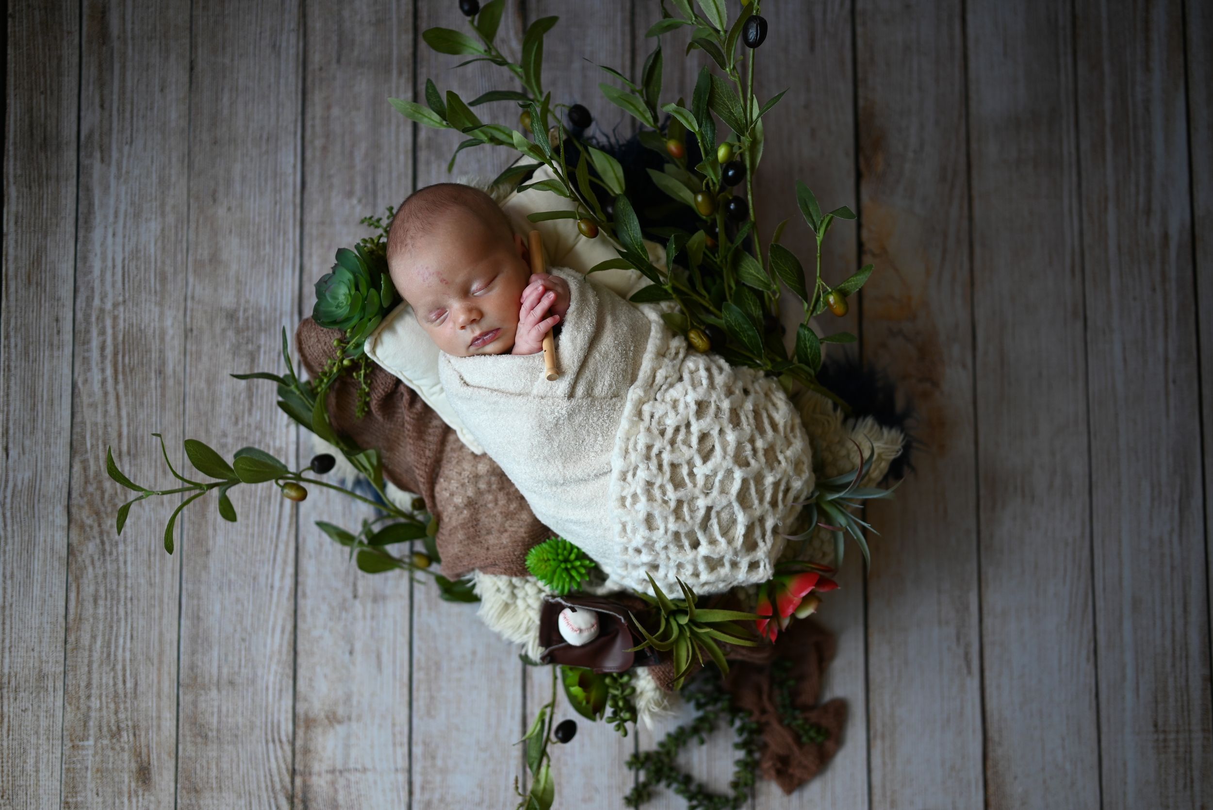 A sleeping newborn swaddled in white is nestled among greenery and flowers on a rustic wooden floor.