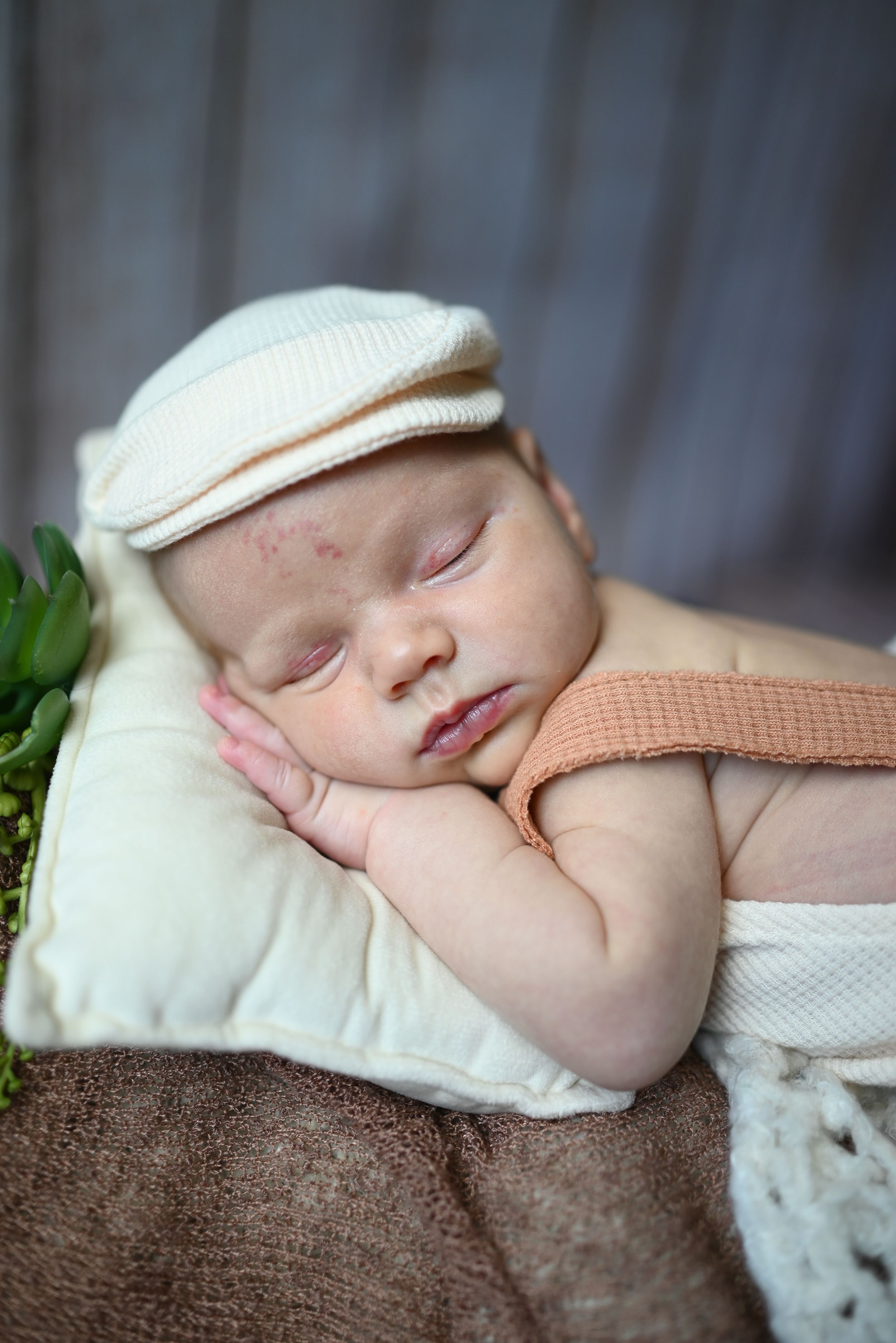 Sleeping newborn wearing white cap and peach outfit rests peacefully on brown blanket with decorative greenery.