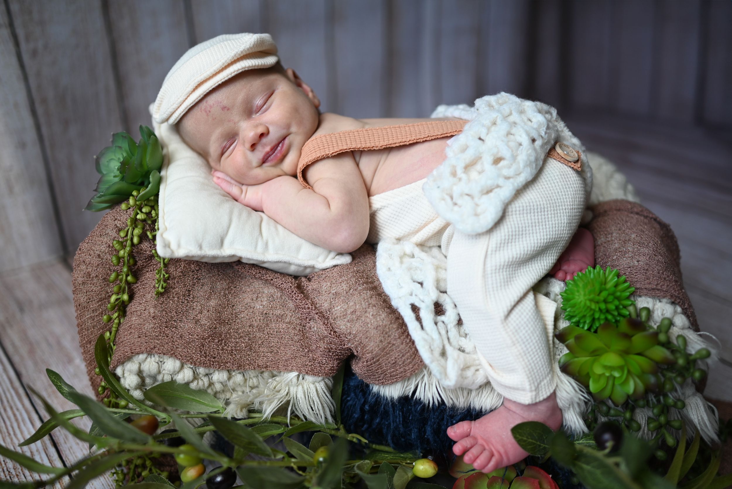 Sleeping newborn baby wearing white outfit rests on brown blanket surrounded by green foliage in studio setting.