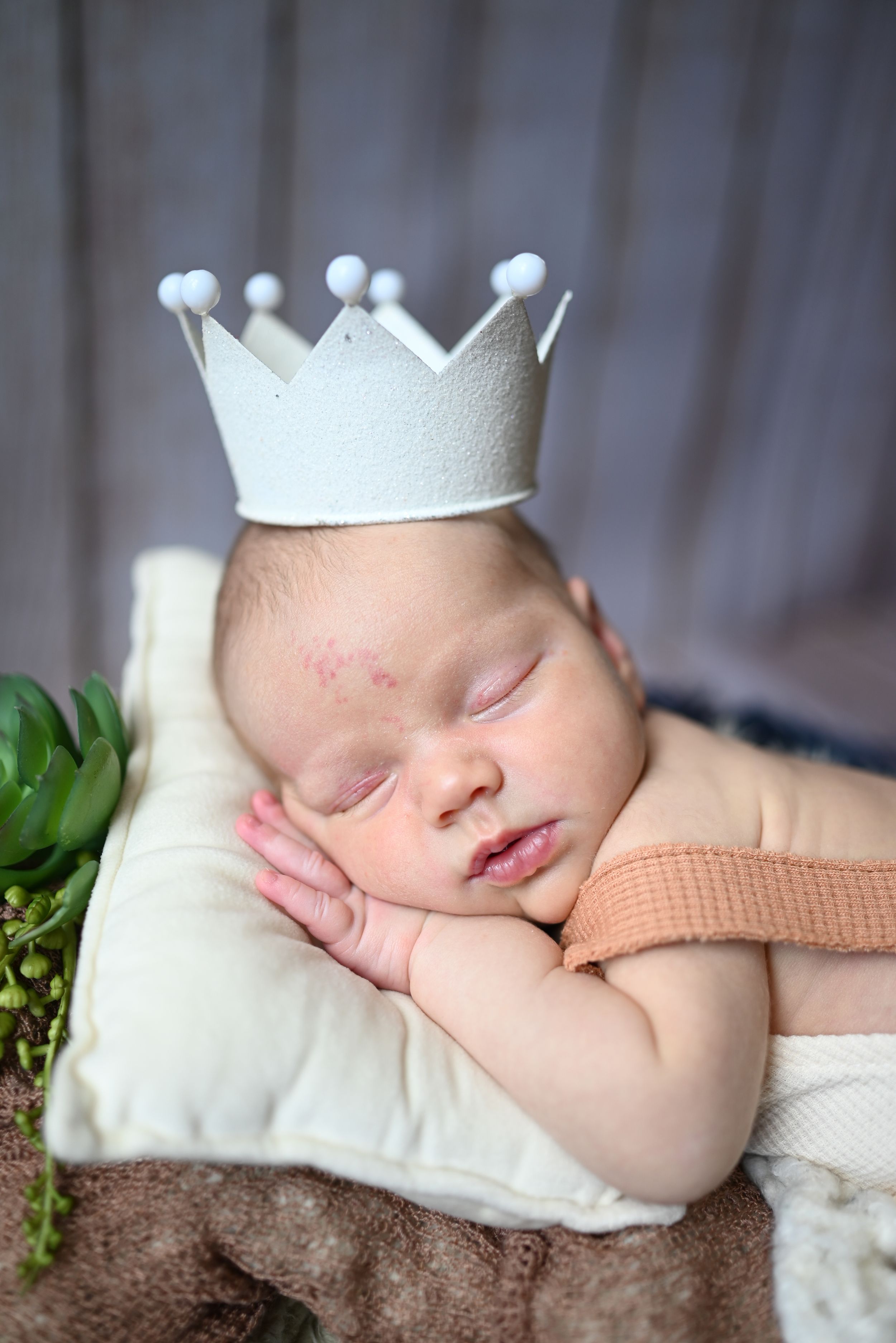 A sleeping newborn wearing a white felt crown rests peacefully on a cream pillow with greenery accents.