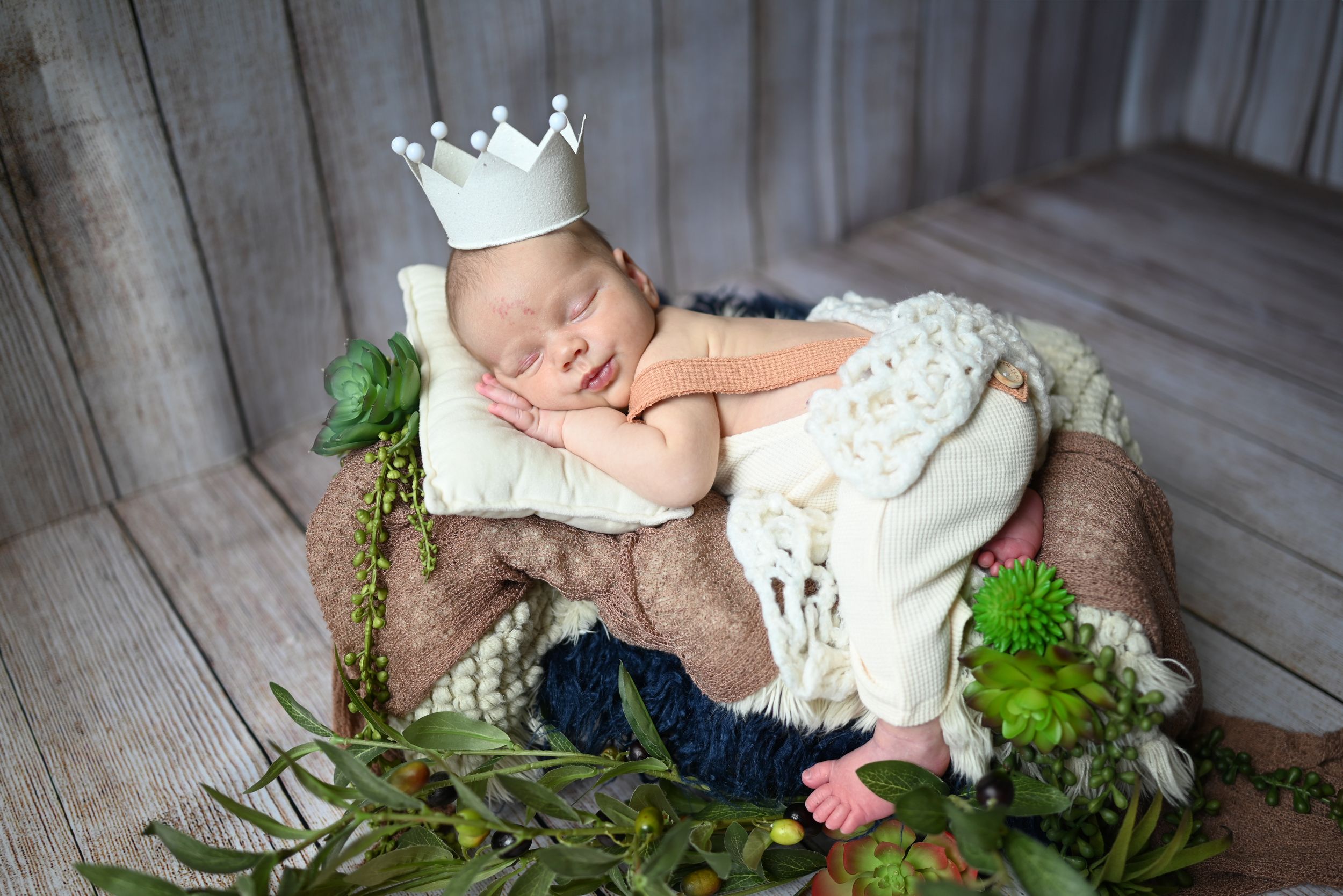 Sleeping newborn wearing white crown and outfit rests on rustic wooden prop with green foliage accents.