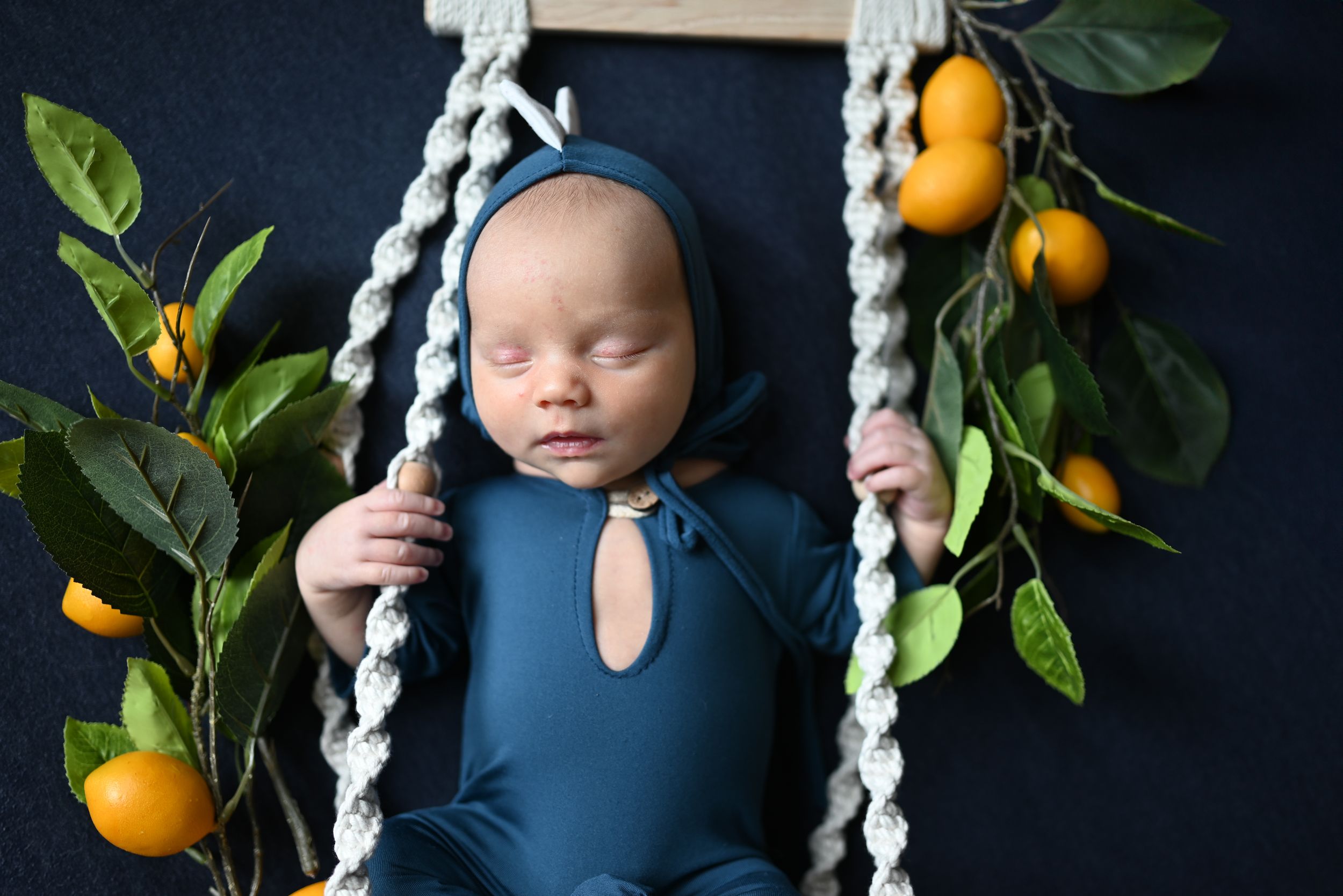 A sleeping infant in a navy blue outfit lies on a beaded swing with citrus branches and dark background.