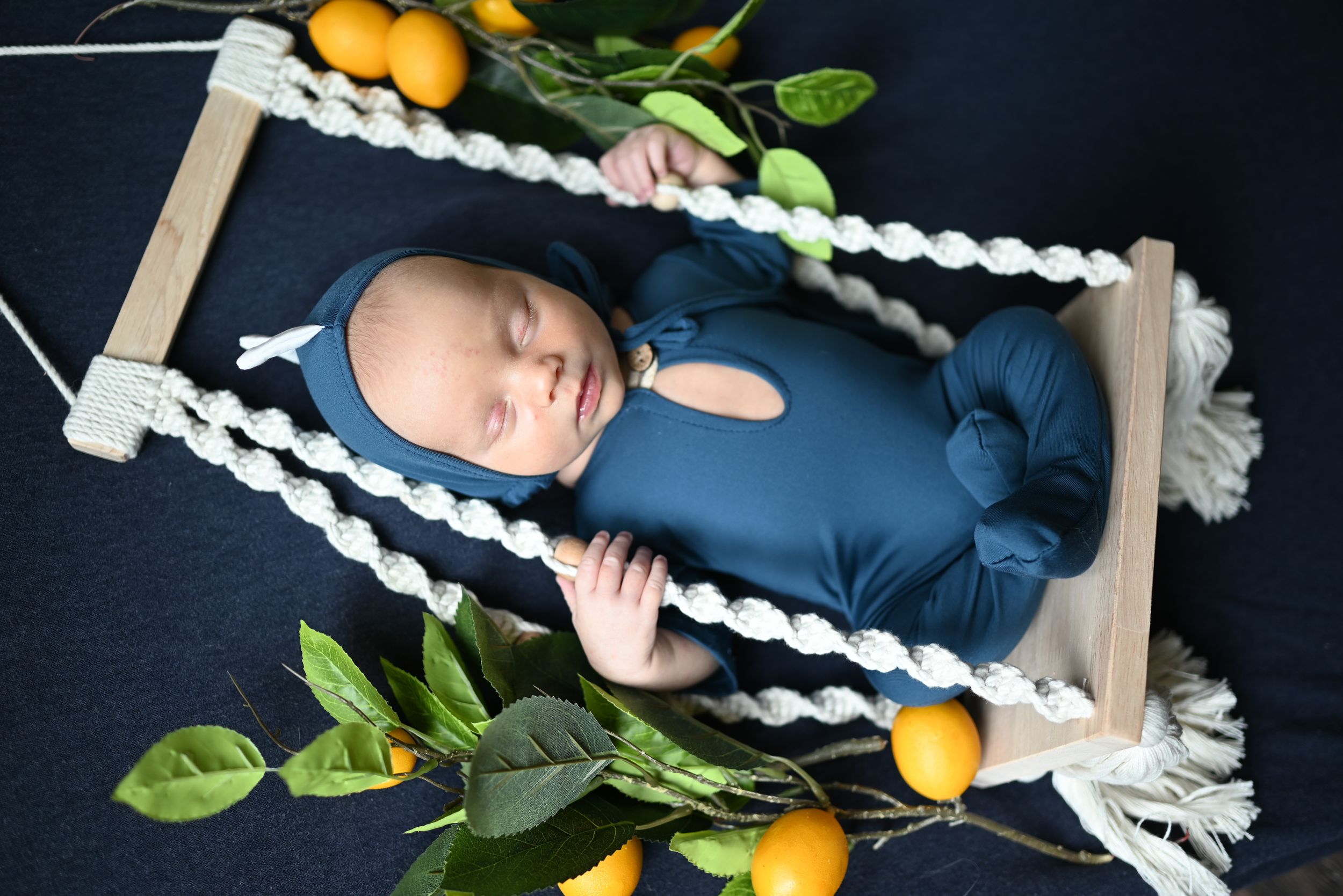 Sleeping baby in navy outfit lies on rope swing surrounded by citrus fruits and green leaves.