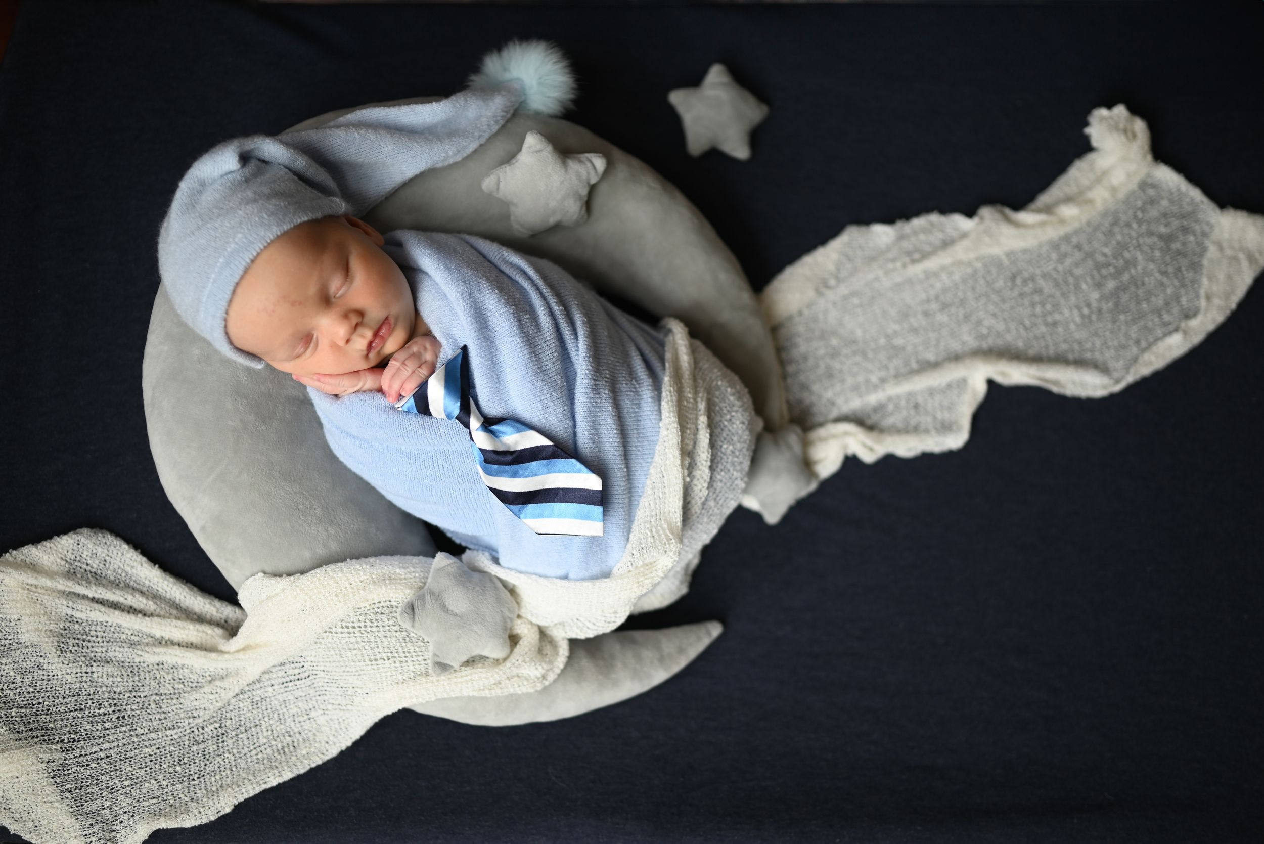 Peaceful newborn in light blue outfit rests on curved gray pillow with flowing fabric backdrop.