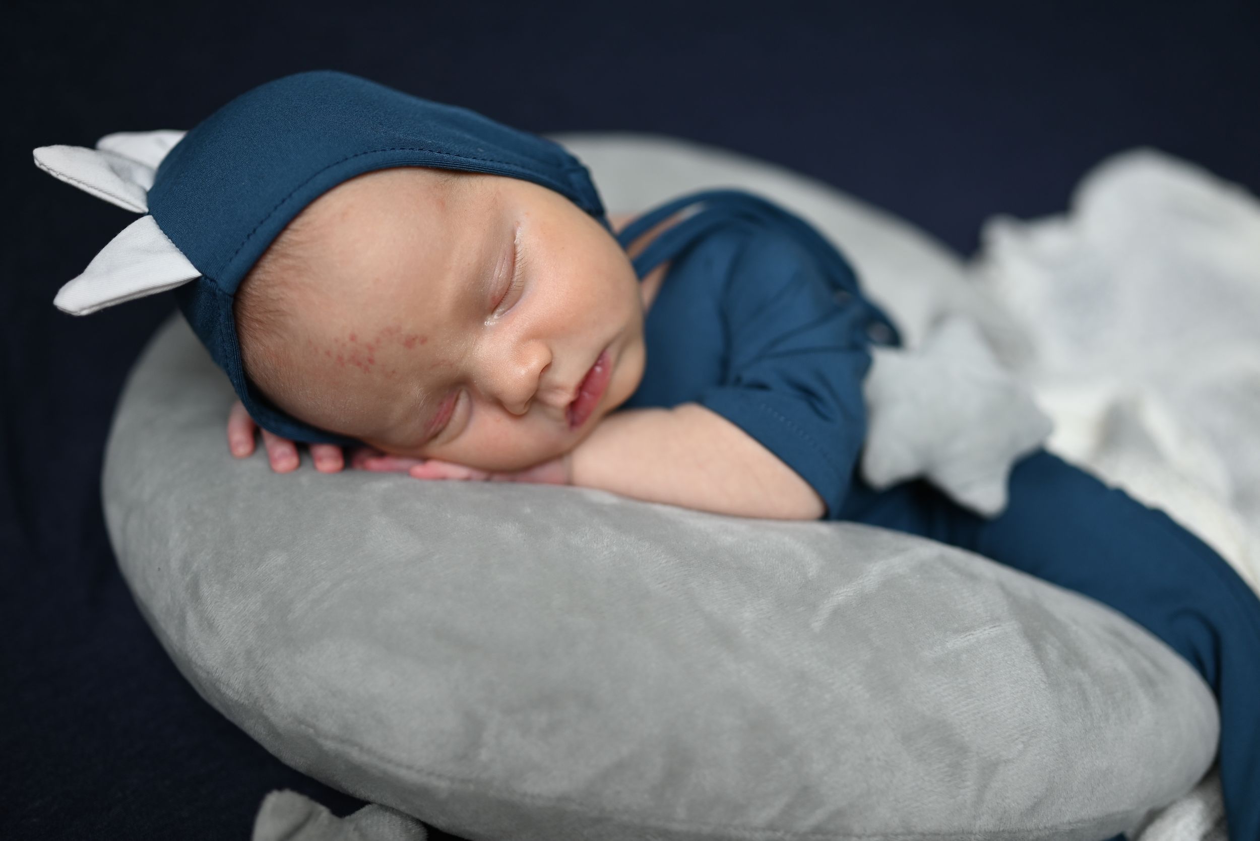A sleeping newborn wearing a navy blue outfit rests peacefully on a soft gray nursing pillow.