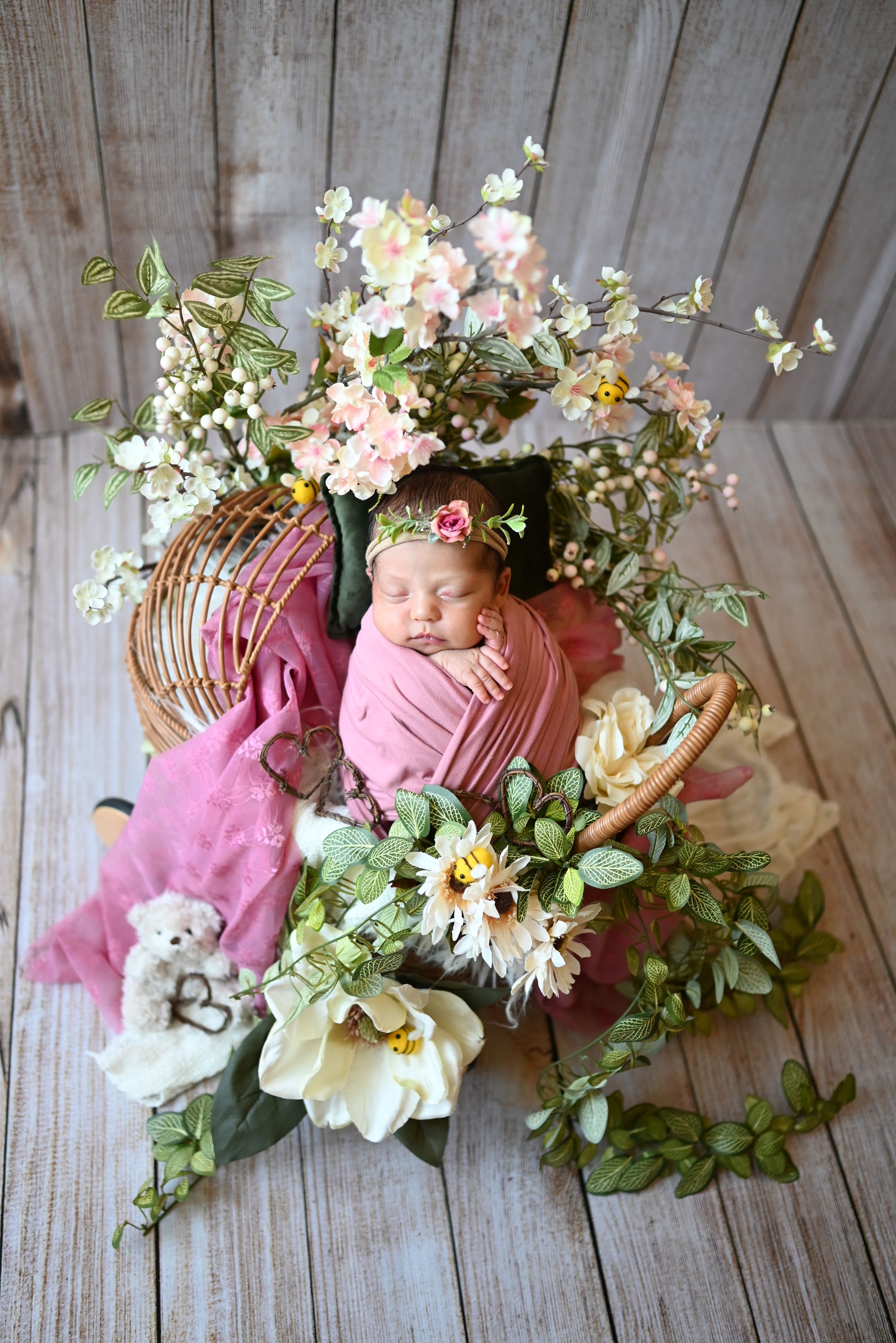 A newborn nestled in pink fabric surrounded by spring flowers and greenery on rustic wooden floor.