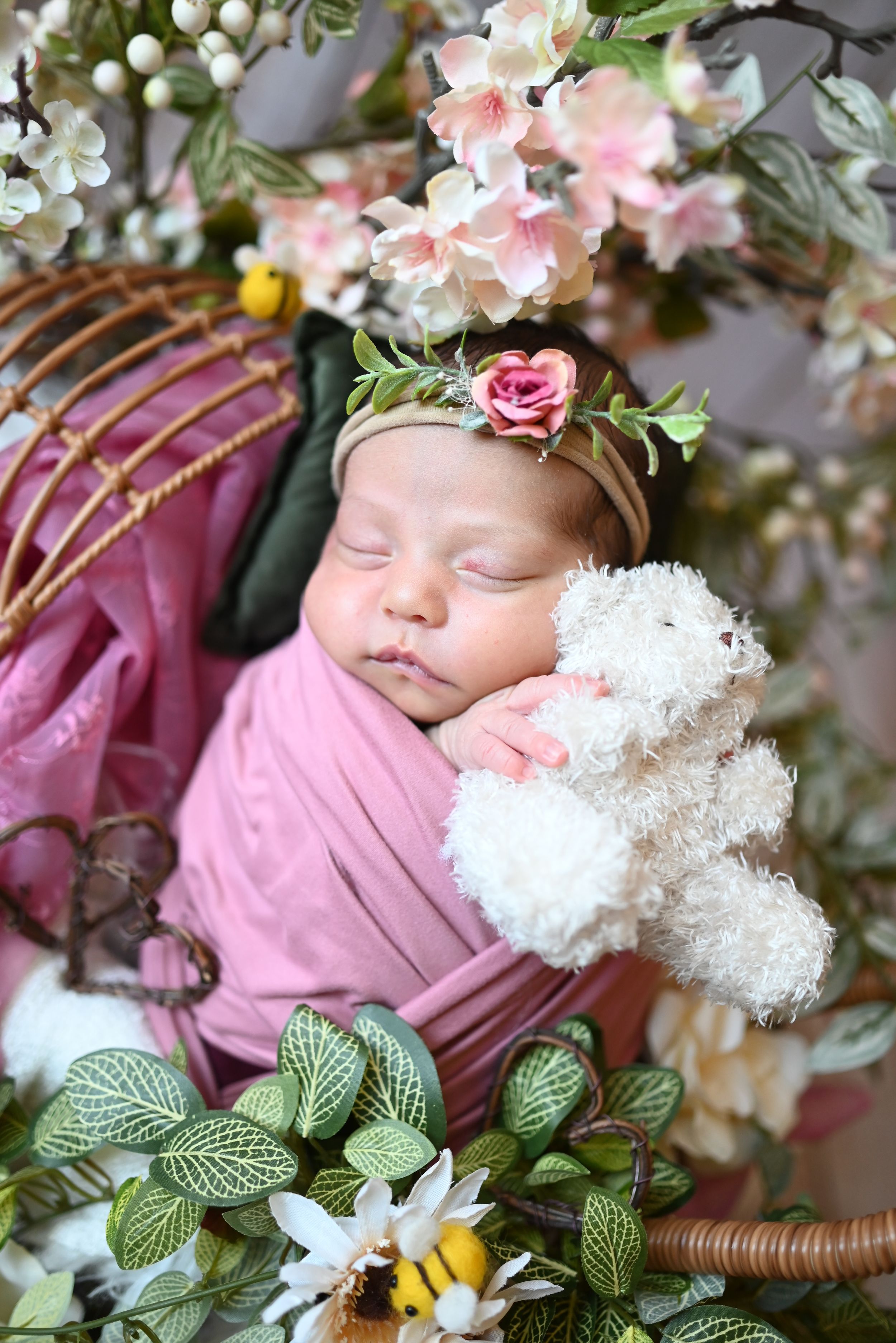 A sleeping infant swaddled in pink with a delicate floral crown nestled among spring blossoms and greenery.