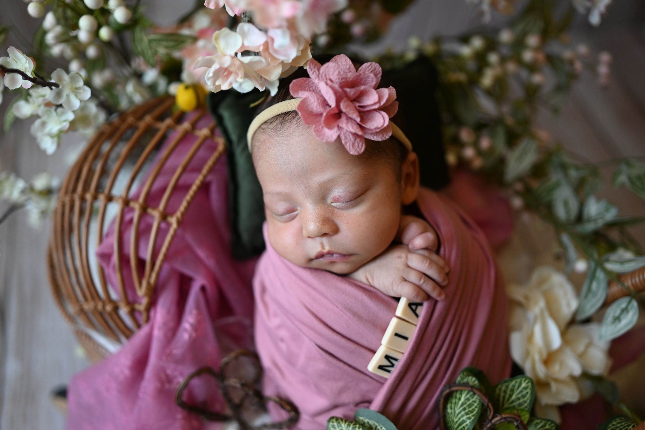 Sleeping newborn swaddled in pink fabric surrounded by delicate pink and white flowers in a rustic basket setting.