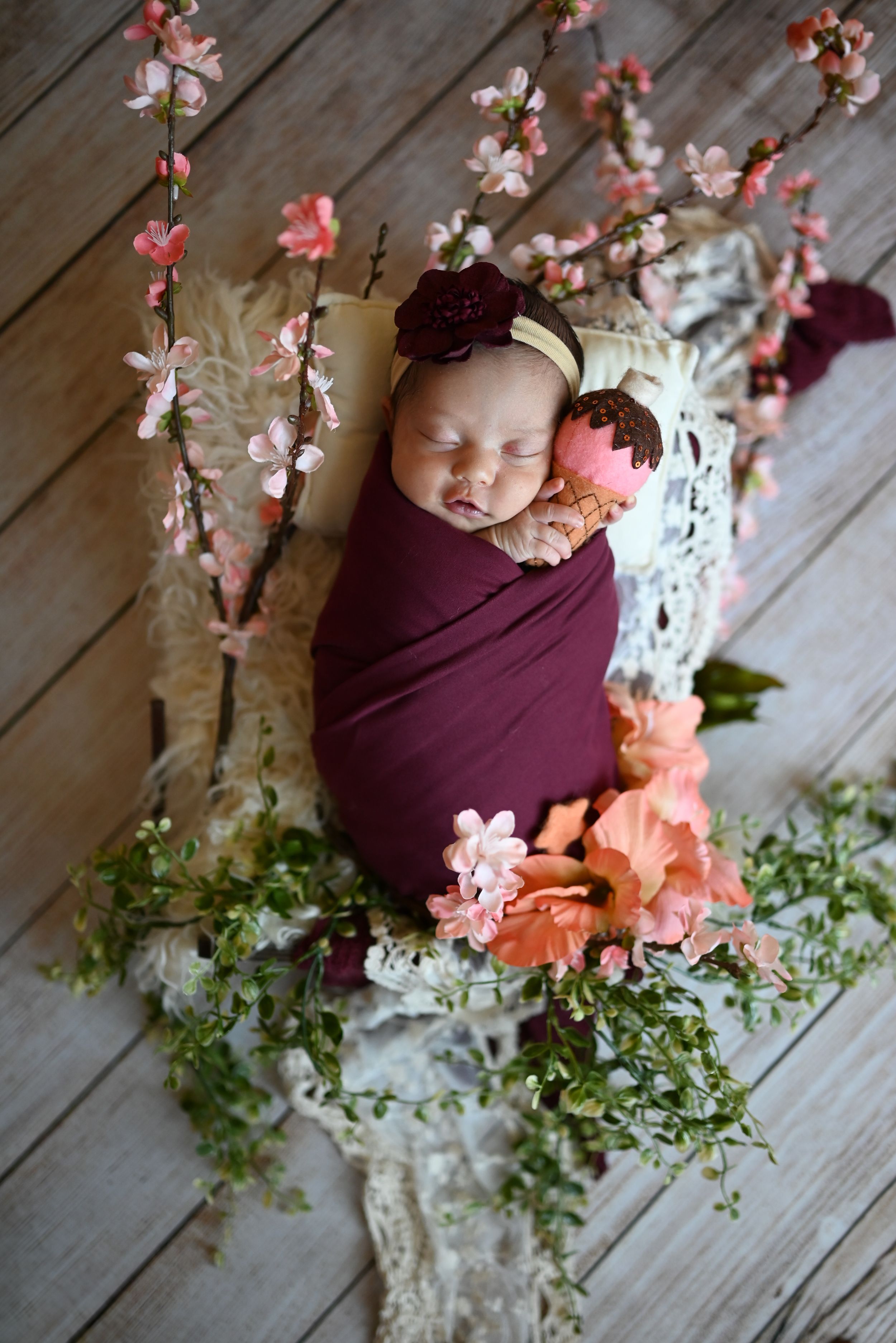 Sleeping newborn wrapped in burgundy fabric surrounded by delicate cherry blossom branches and greenery on wooden floor.
