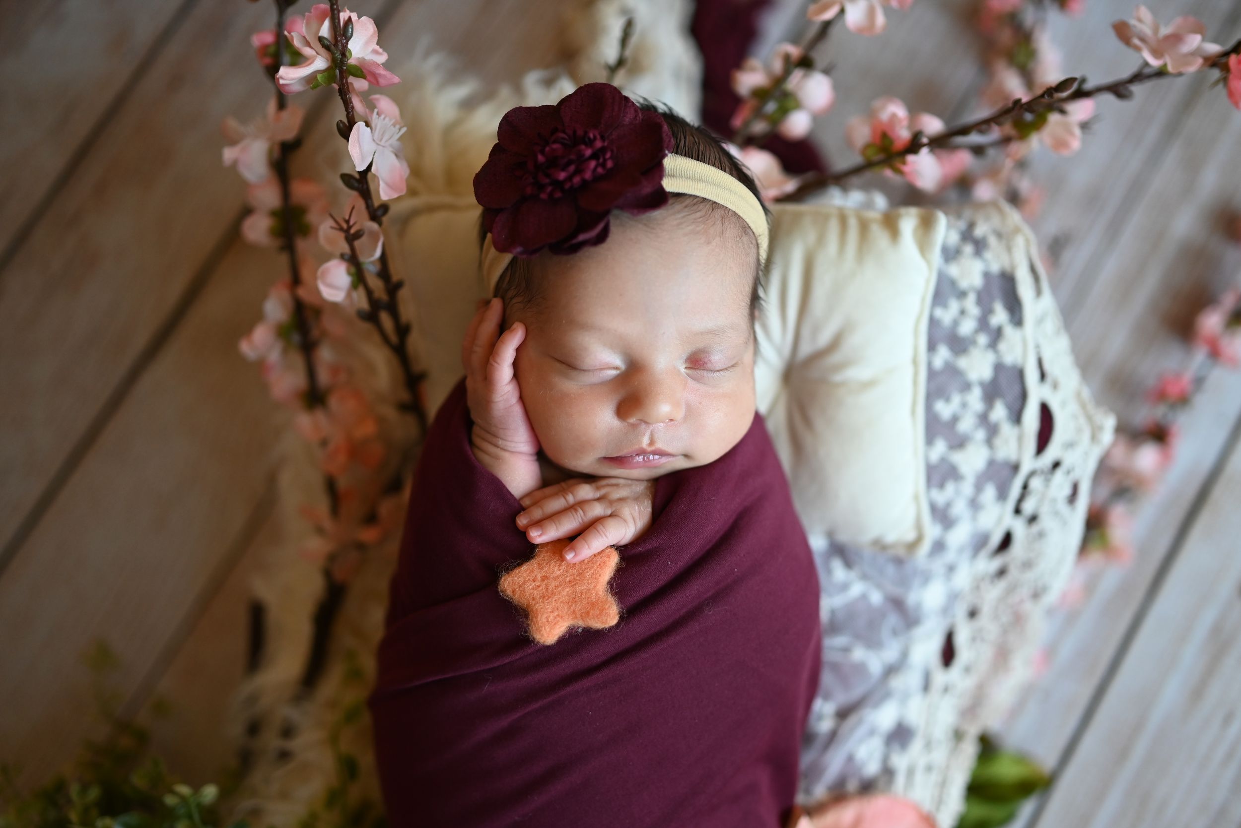 A baby wrapped in maroon fabric sleeps peacefully among delicate pink cherry blossoms and silver birch branches.