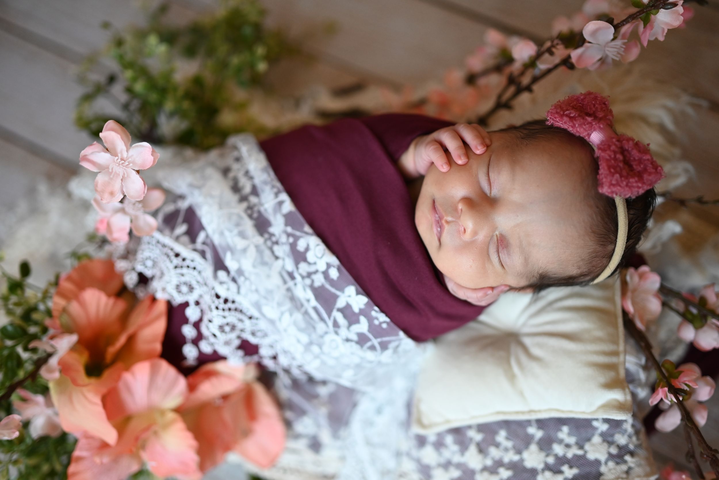 A sleeping newborn wrapped in burgundy and lace swaddle with pink floral decorations and headband.
