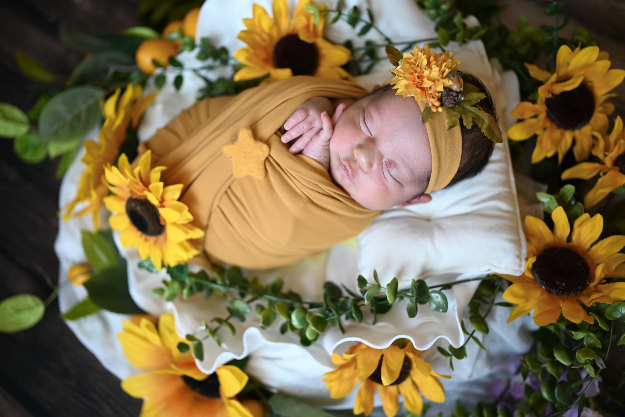 Peaceful newborn swaddled in mustard yellow wrap nestled among bright sunflowers and greenery on white blanket.