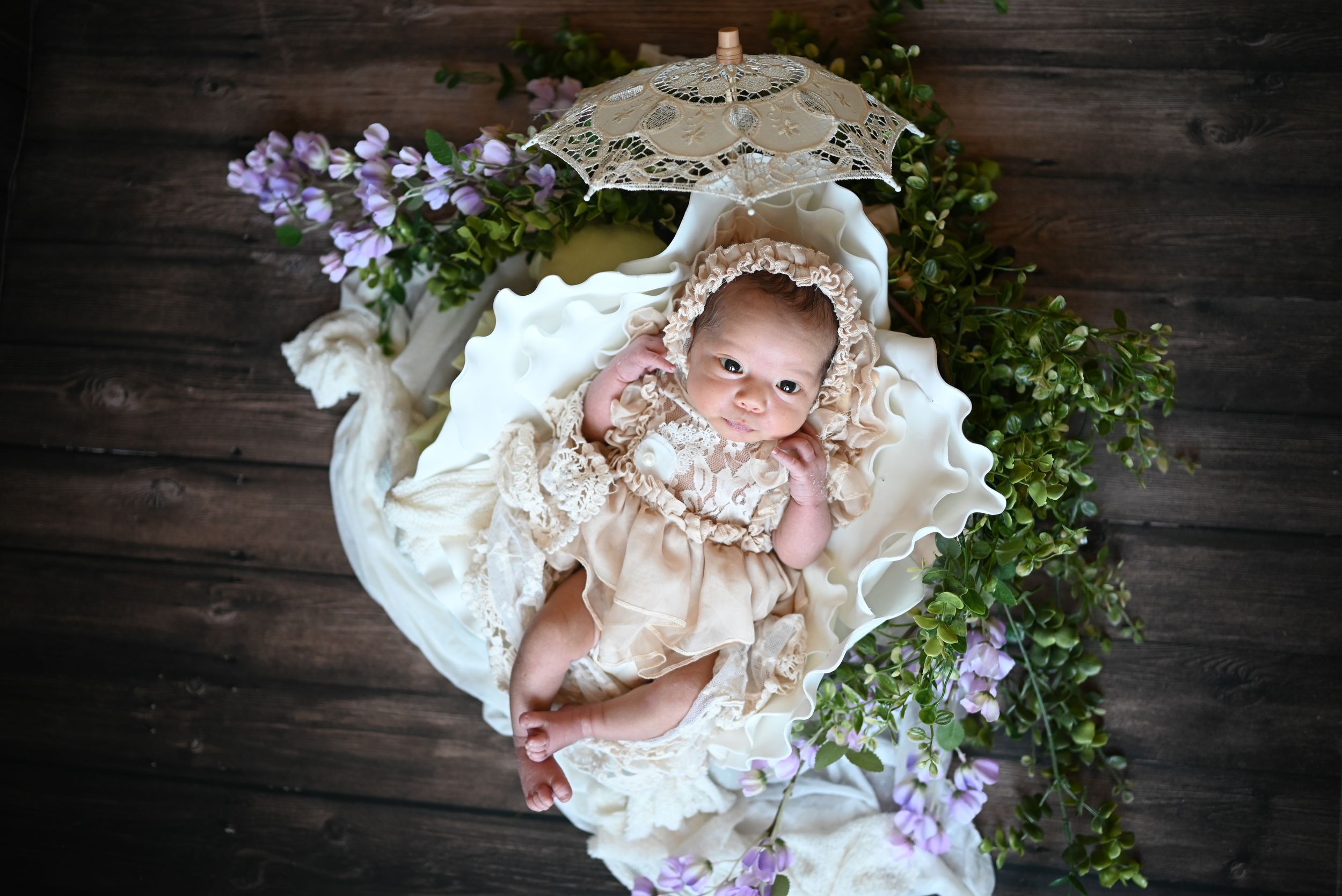 Pale pink silk dress with lace trim lies artfully arranged among delicate purple flowers on dark wooden surface.