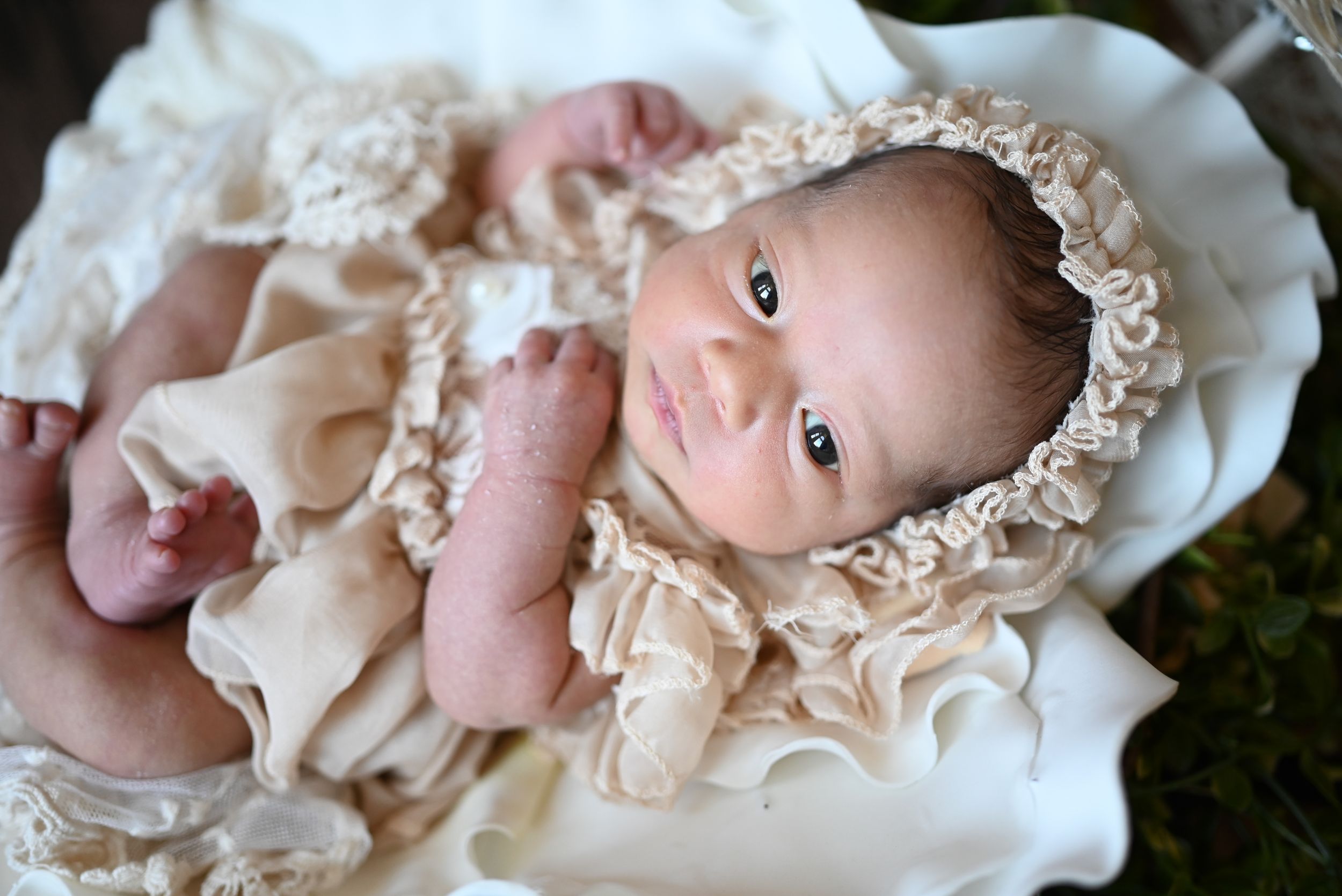 A tiny baby wearing a delicate cream colored bonnet and dress lays on white fabric.