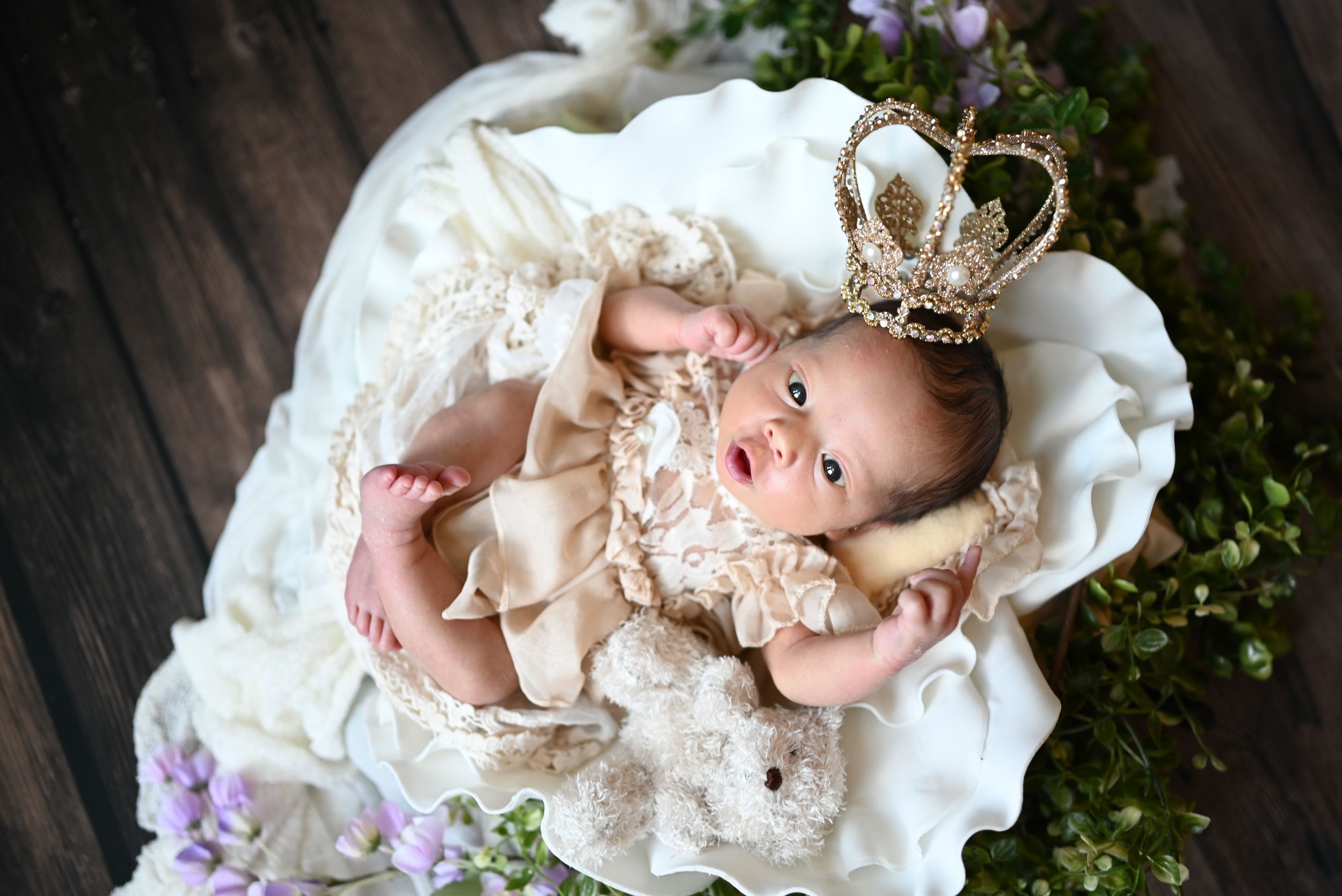 A small baby wearing lace dress and crown lies on white fabric surrounded by purple flowers on wooden background.