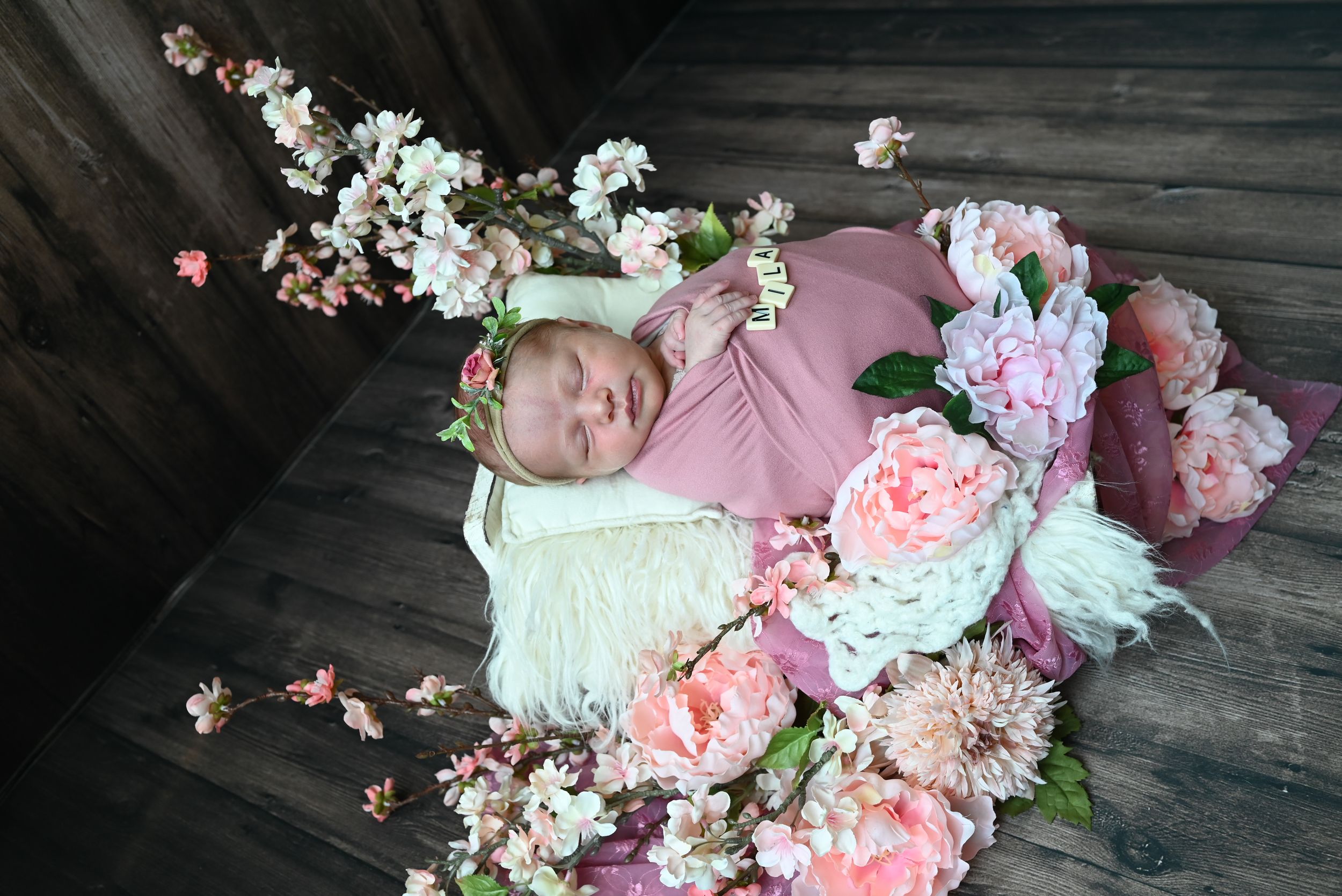 Sleeping baby in pink outfit lies on white blanket surrounded by delicate cherry blossoms and pink floral arrangements.