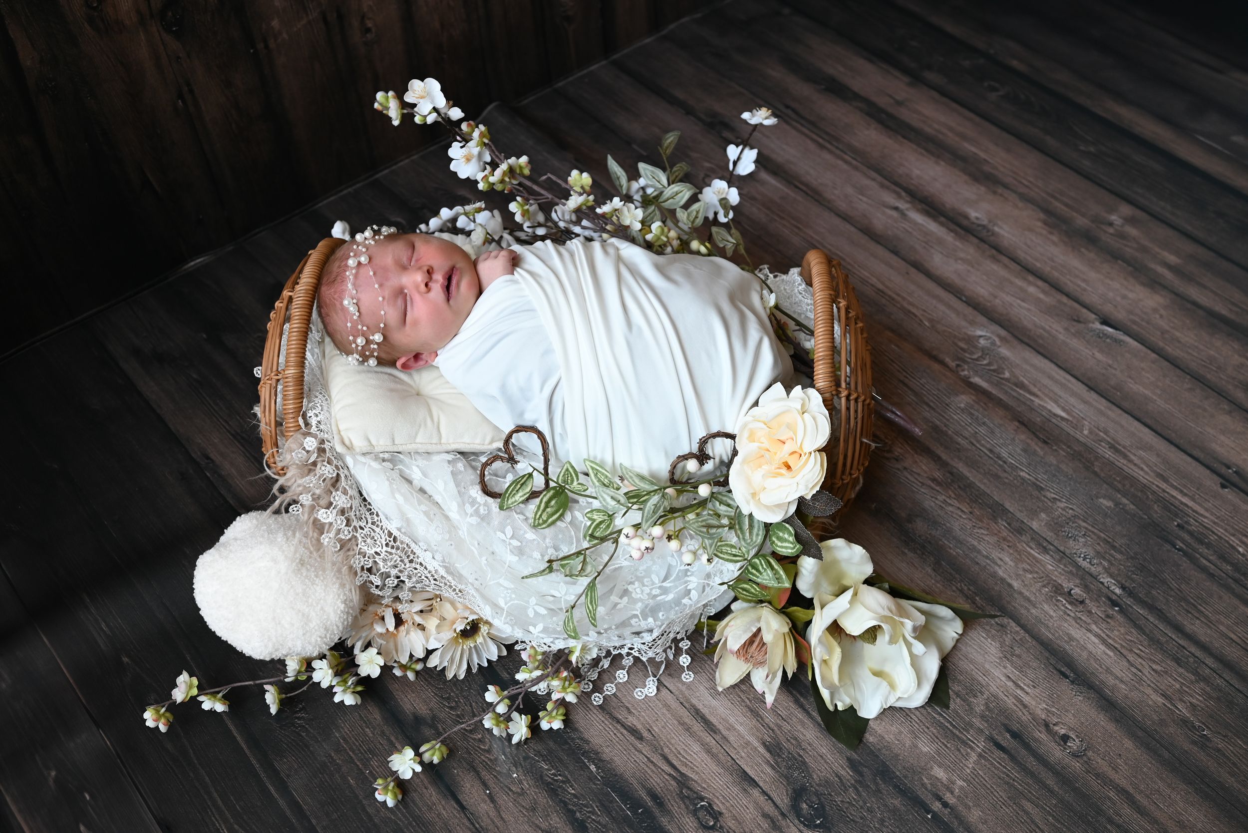 Newborn wrapped in white swaddle lying in wooden basket with flowers and delicate baby's breath on dark wood floor.