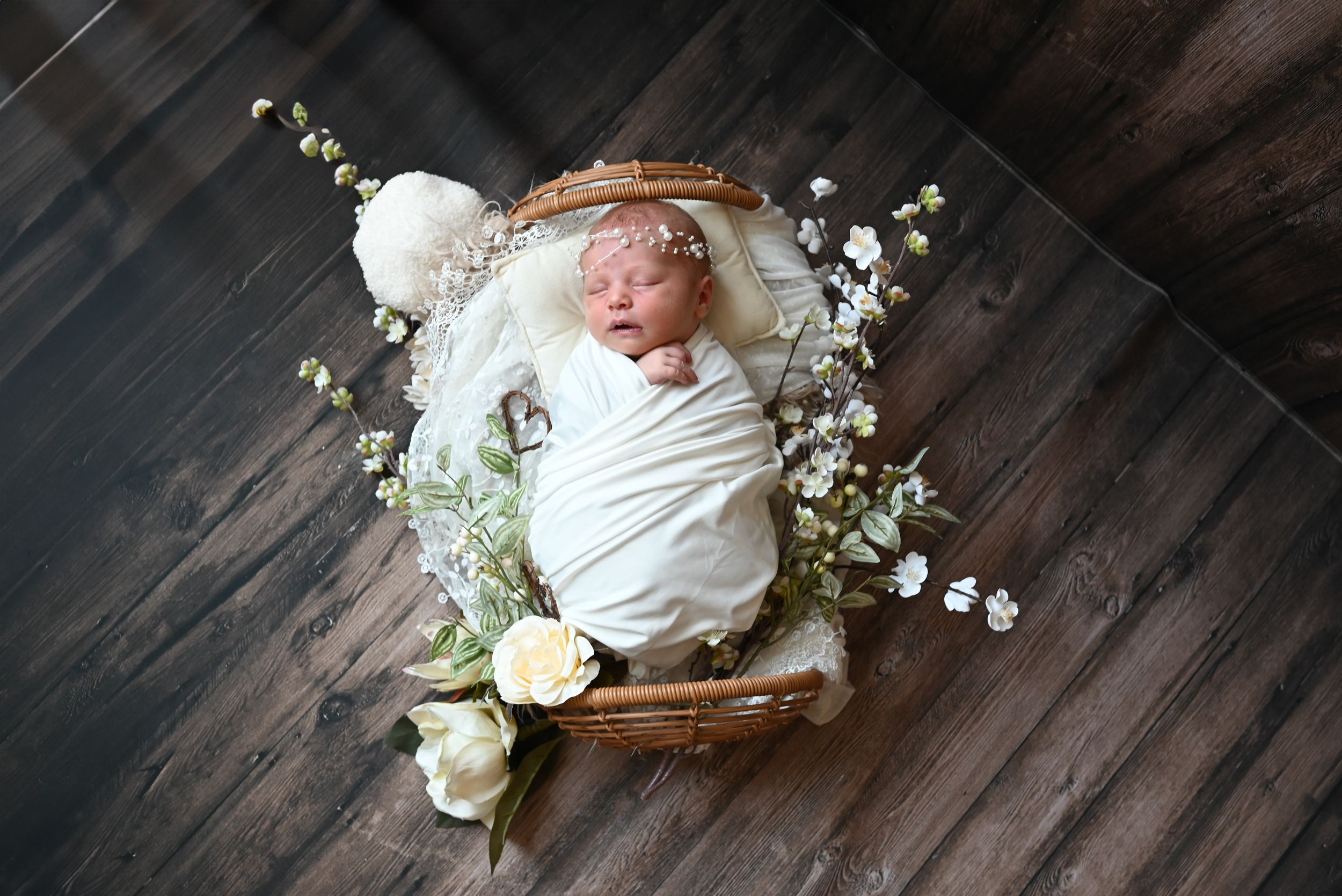 Newborn in white swaddle nestled in rustic wooden bowl with roses and baby's breath on hardwood flooring.