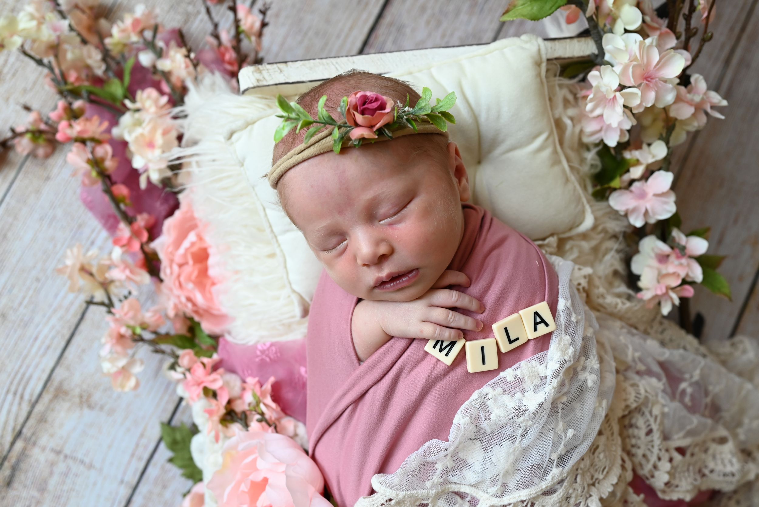 Newborn wrapped in pink fabric sleeps peacefully surrounded by spring blossoms and floral decor on rustic wood floor.