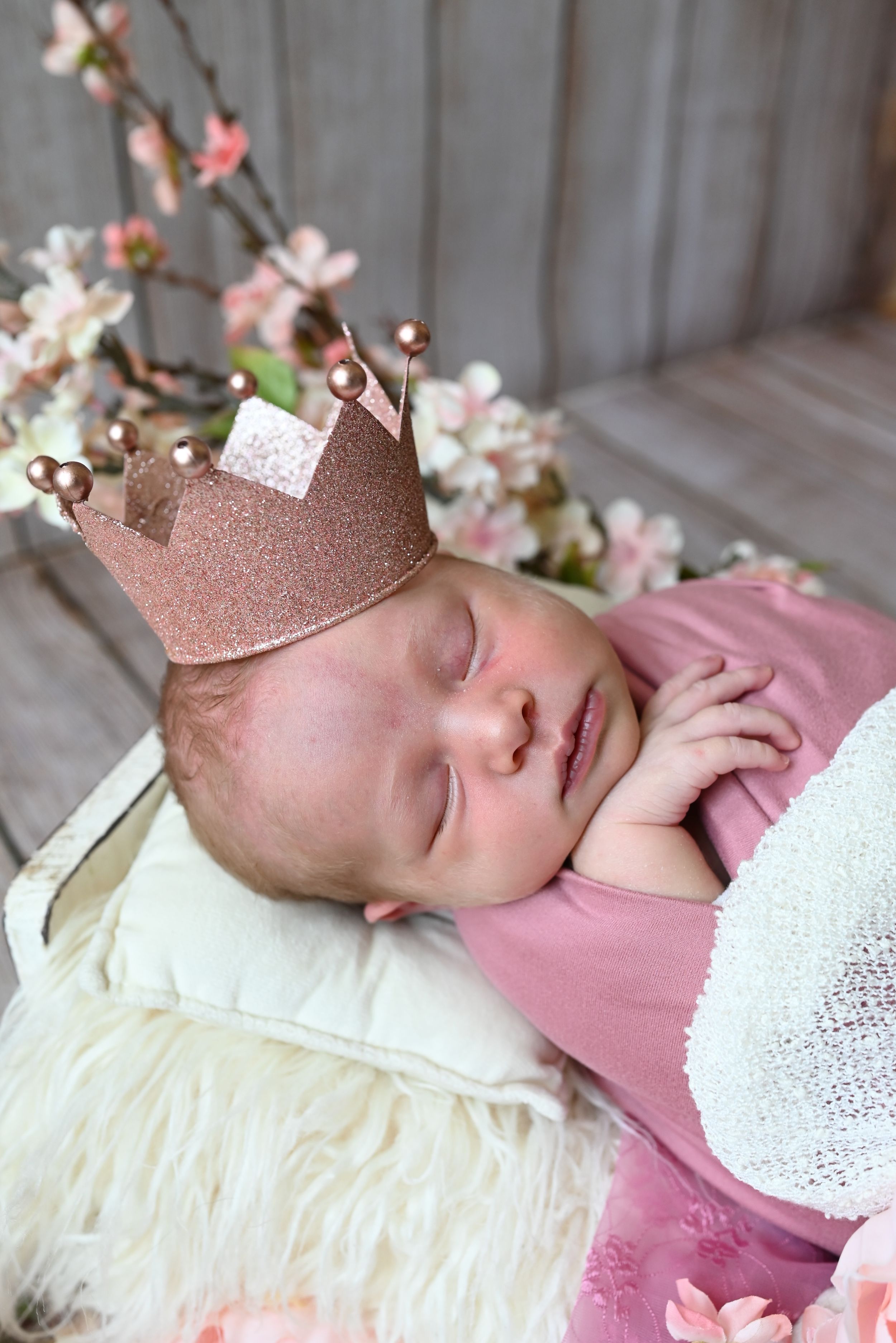 A sleeping baby wearing a rose gold crown and pink outfit lays on white lace bedding with cherry blossoms nearby.