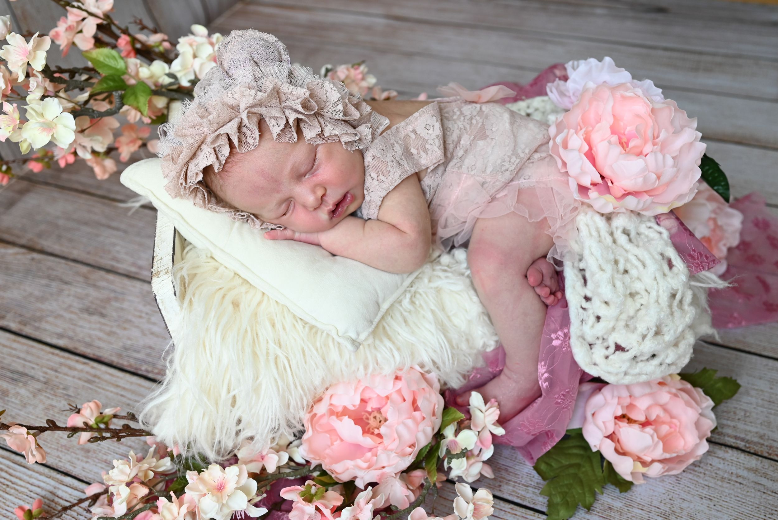 A sleeping baby surrounded by pink and white peonies lies peacefully on rustic wooden floor planks.