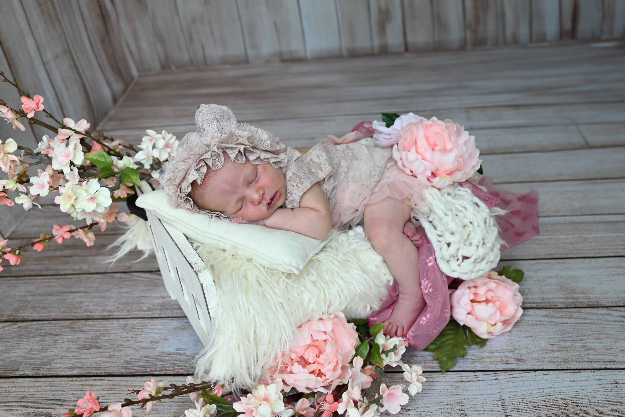 A sleeping infant in cream lace dress lies on rustic wooden floor surrounded by soft pink flowers and cherry blossoms.