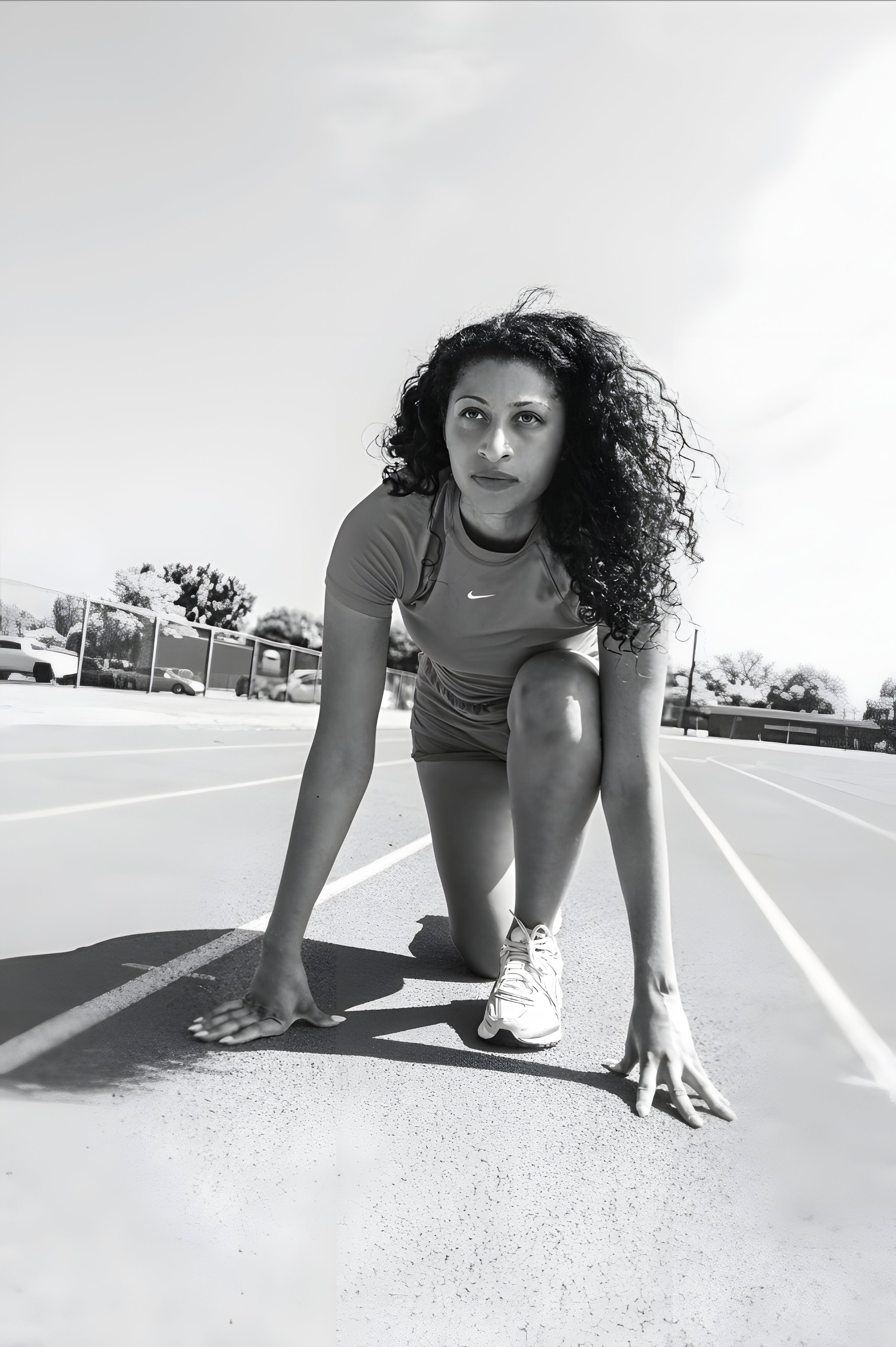 An athlete with curly hair in a Nike shirt assumes a starting position on a running track in black and white photography.