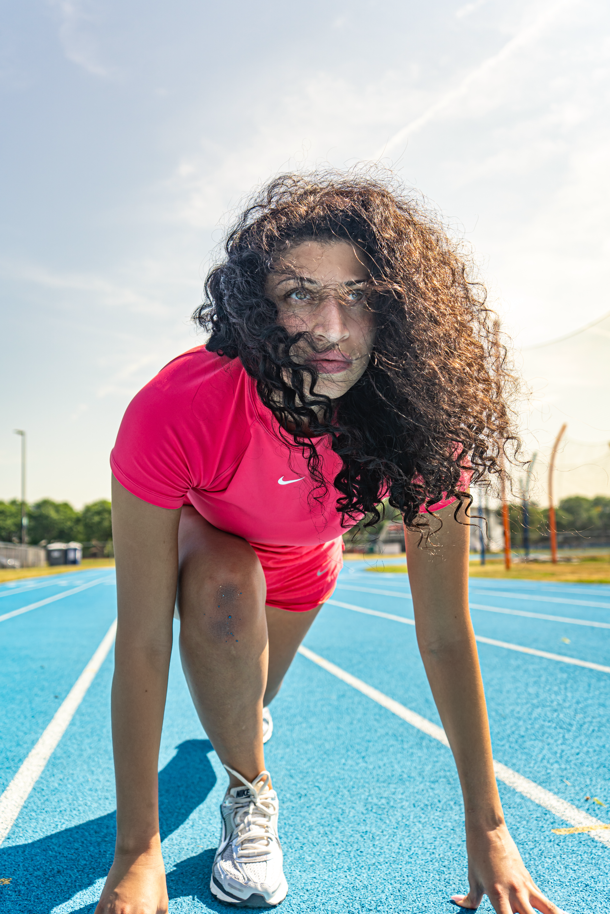 Athlete in red Nike shirt crouches in starting position on a bright blue running track on a sunny day.