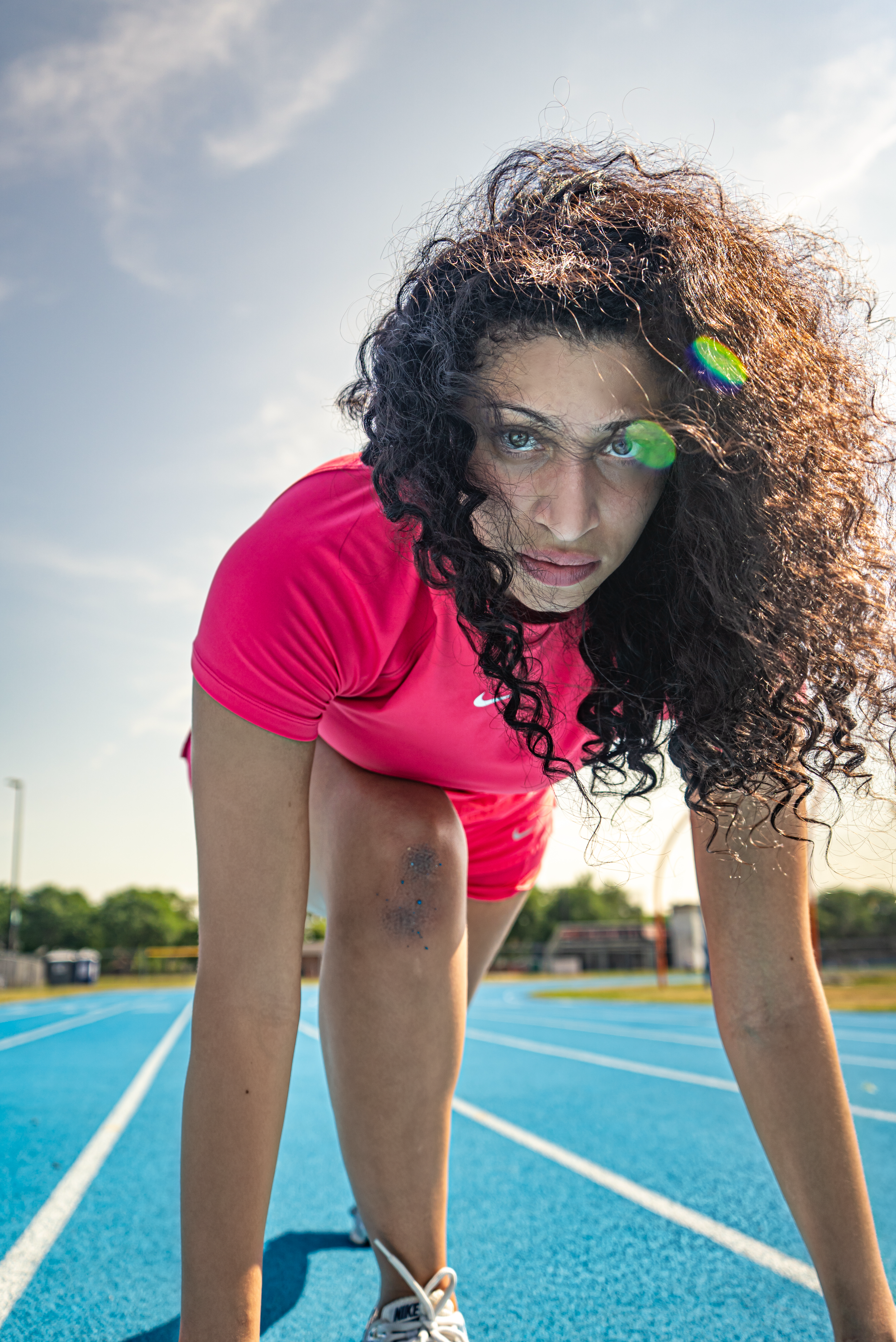 Athlete in pink shirt at starting position on blue running track during sunset.
