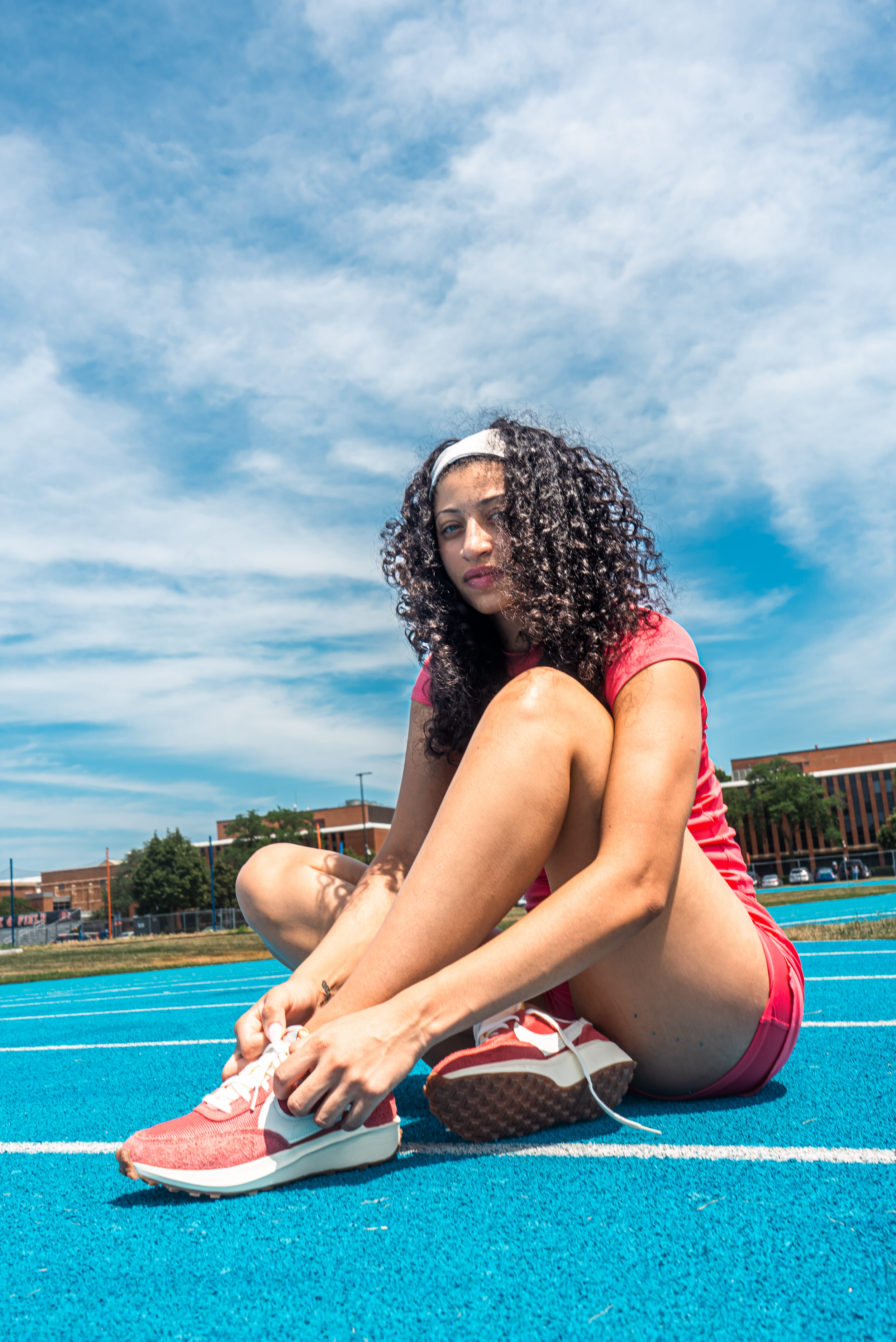 Athlete tying shoelaces while sitting on bright blue running track surface under cloudy summer sky.