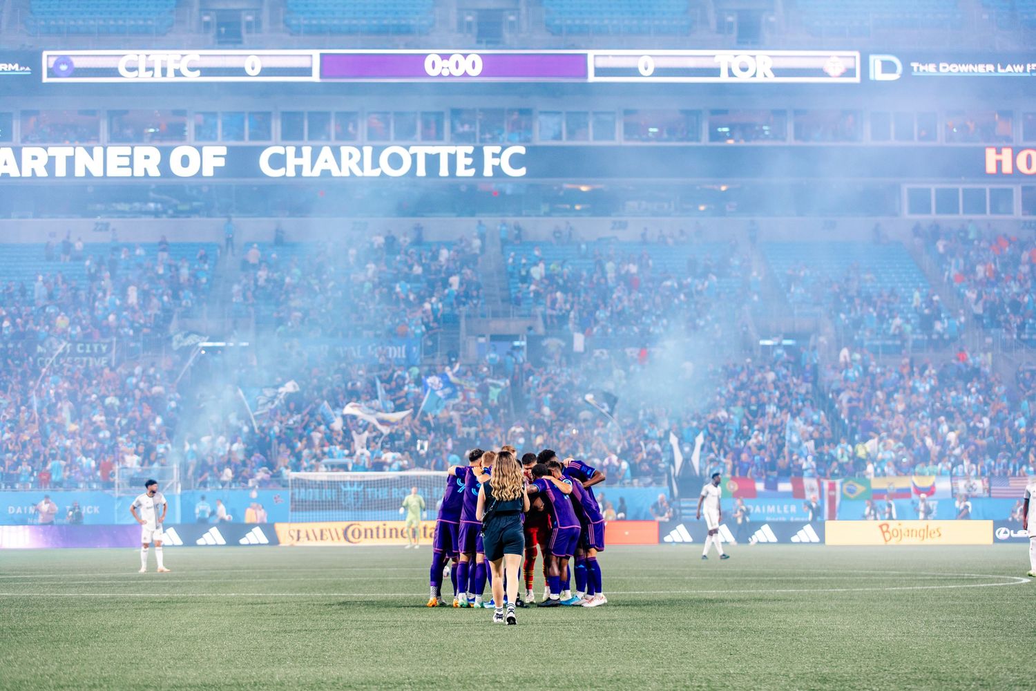 Charlotte FC soccer team huddles in stadium with blue smoke and fans.